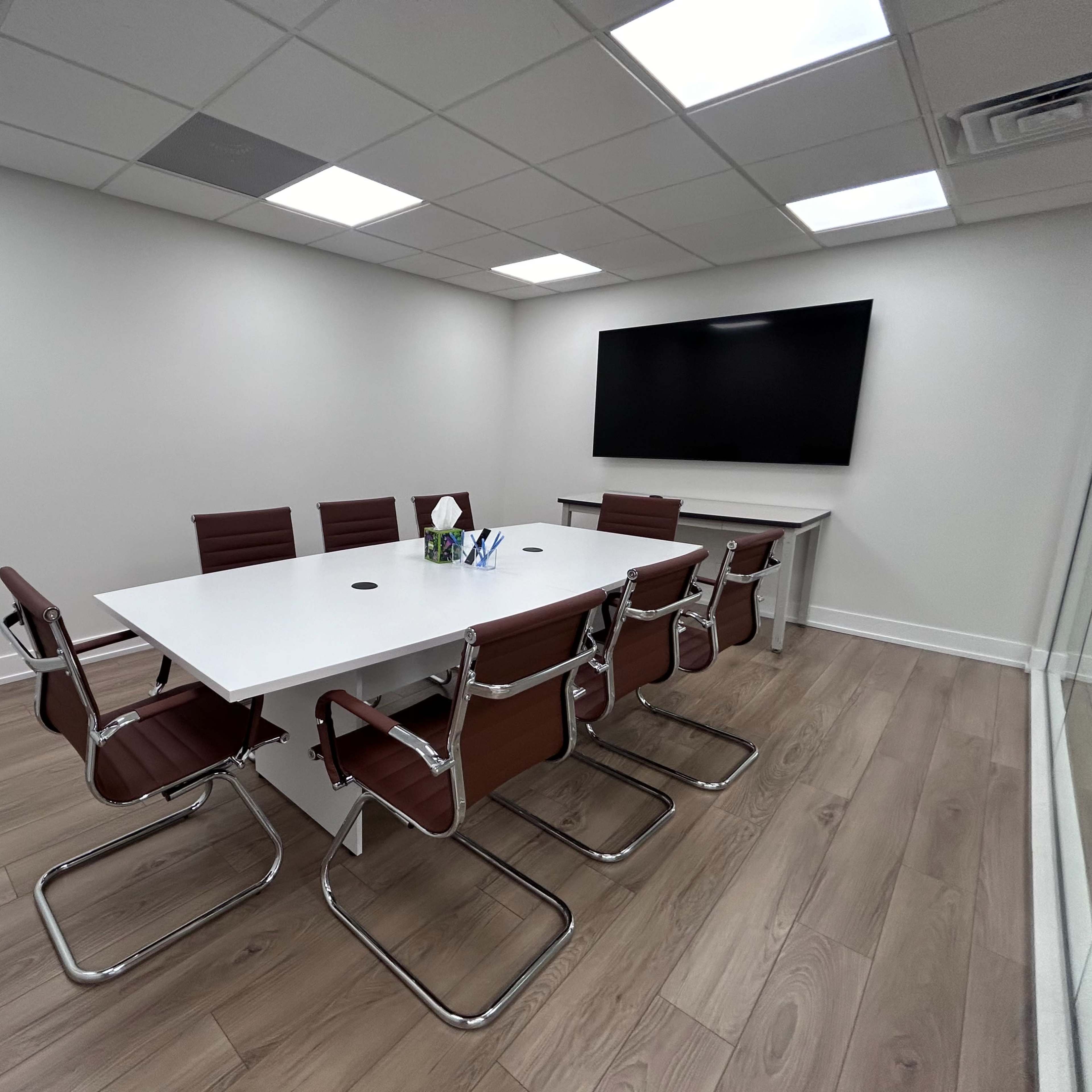 A modern conference room features a white table surrounded by brown leather chairs, with a large screen mounted on the wall.