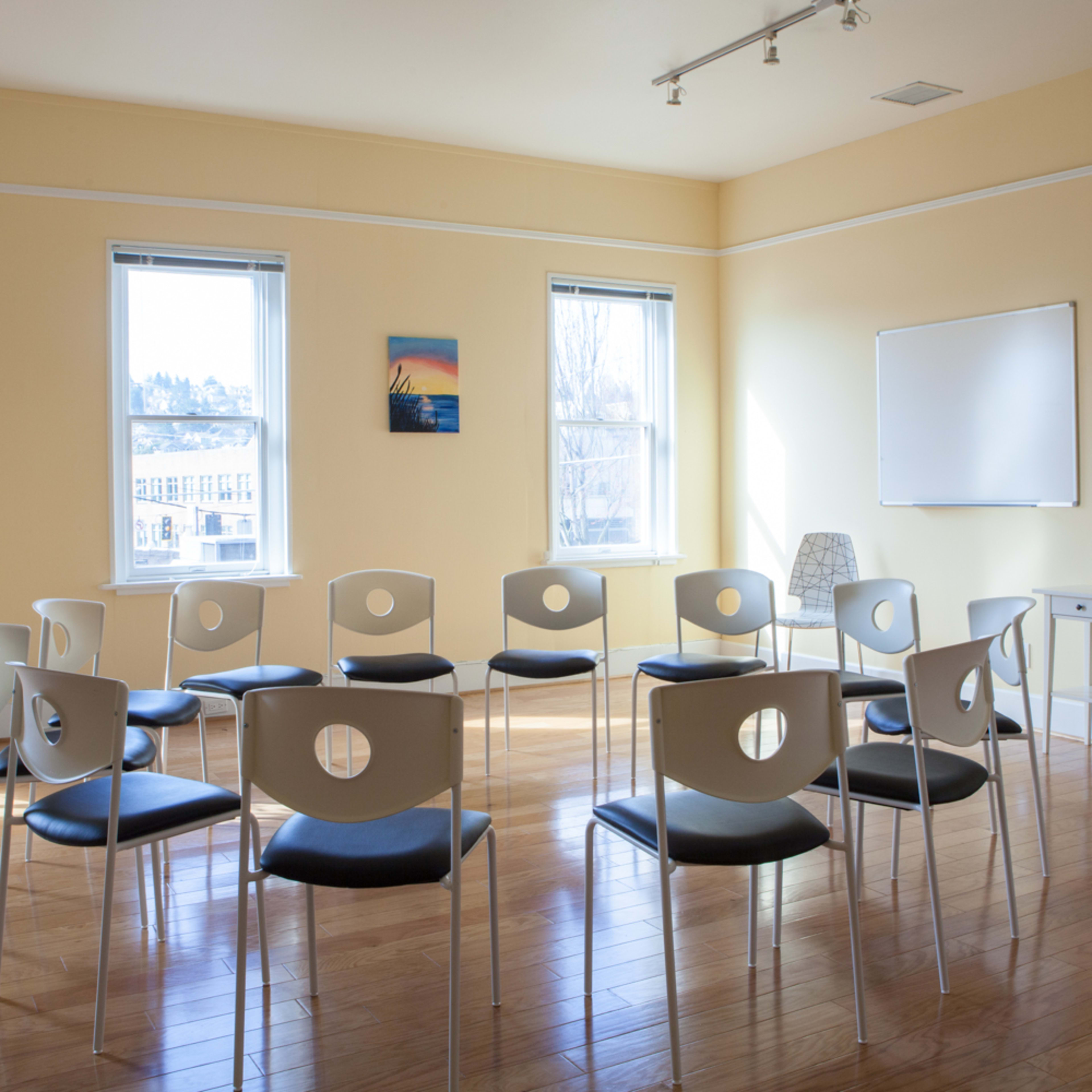 A circle of chairs is arranged in a room with light wood flooring and large windows.