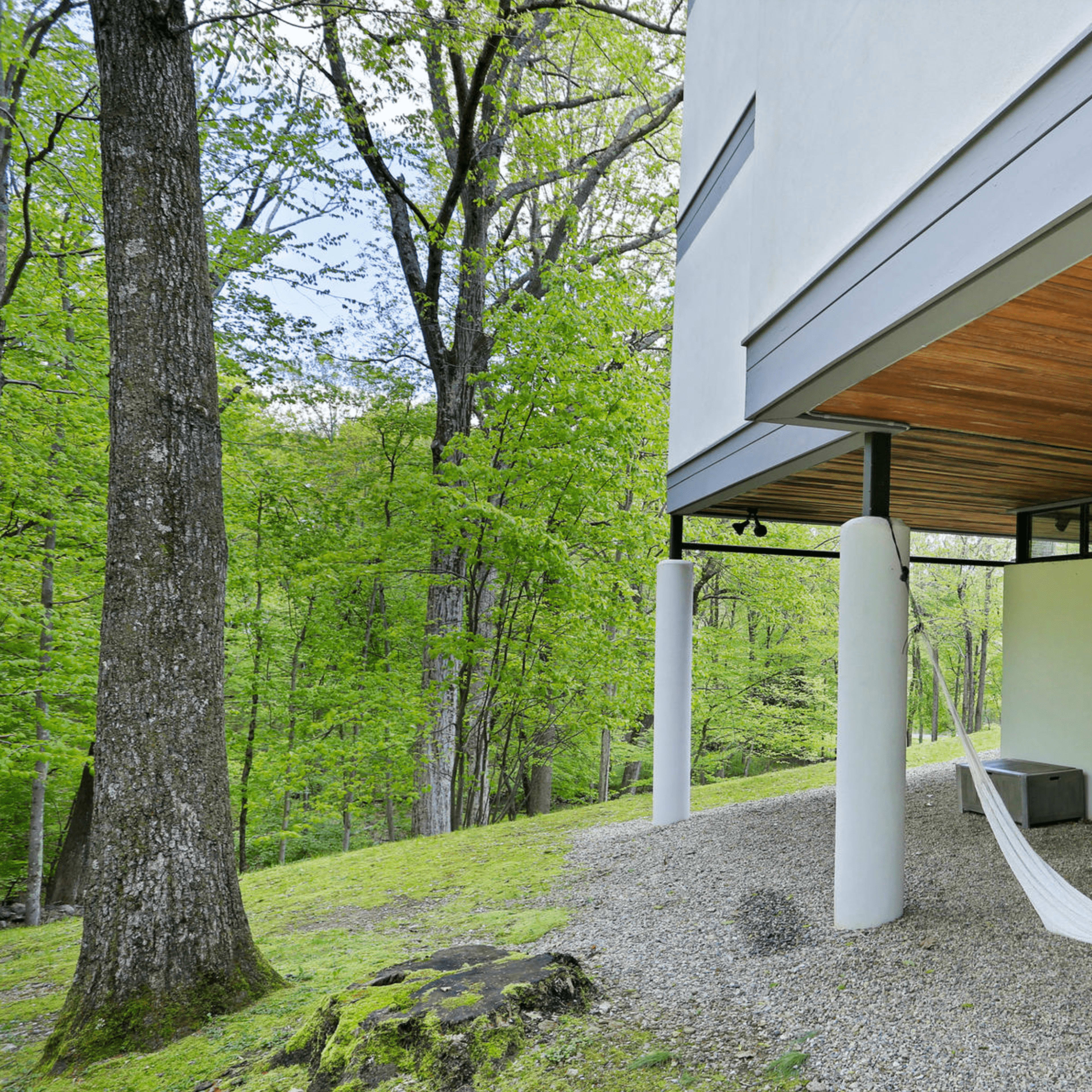A modern house with a wooden overhang features a hammock suspended between two columns, surrounded by green trees and gravel landscaping.