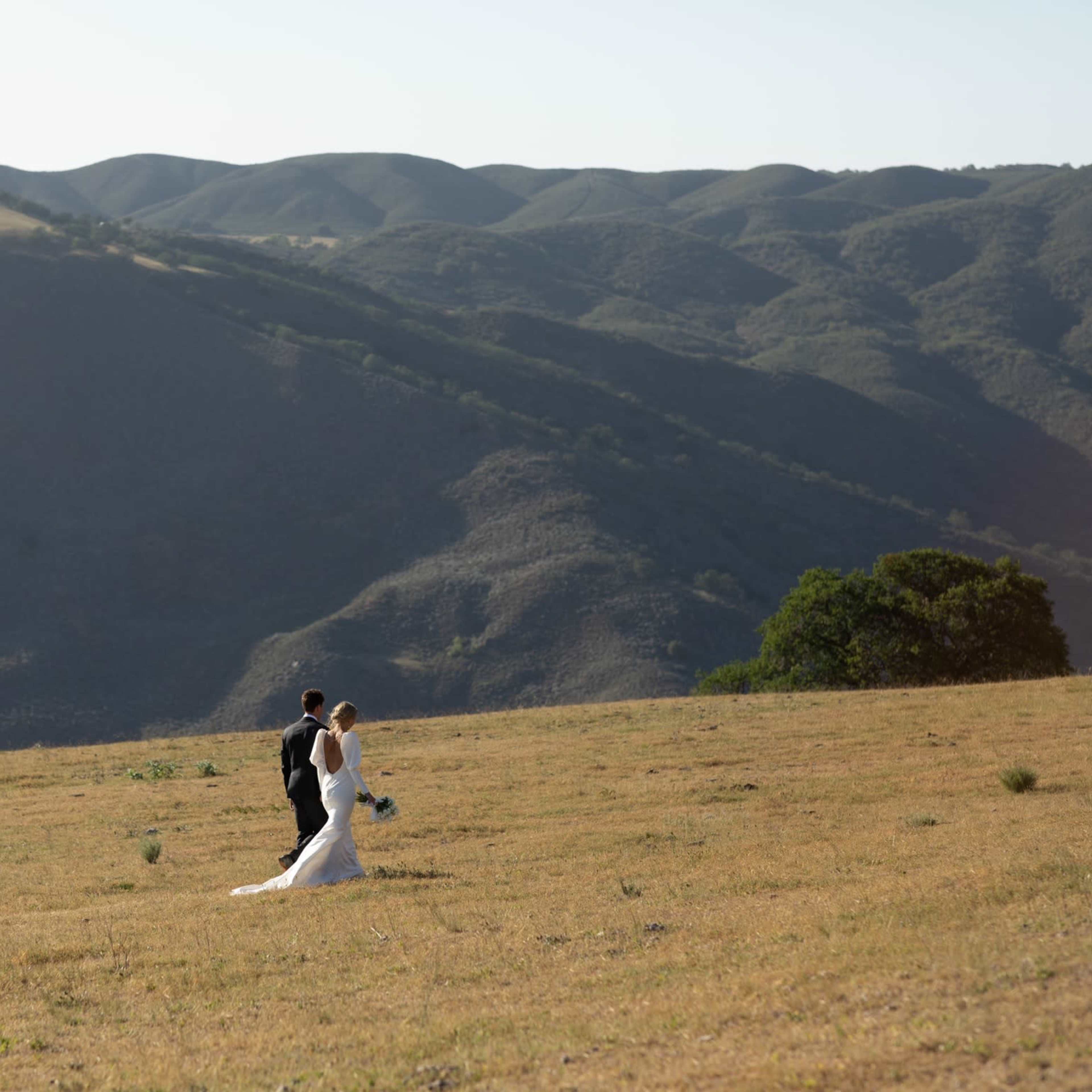 A couple walks together across a grassy field with rolling hills in the background.