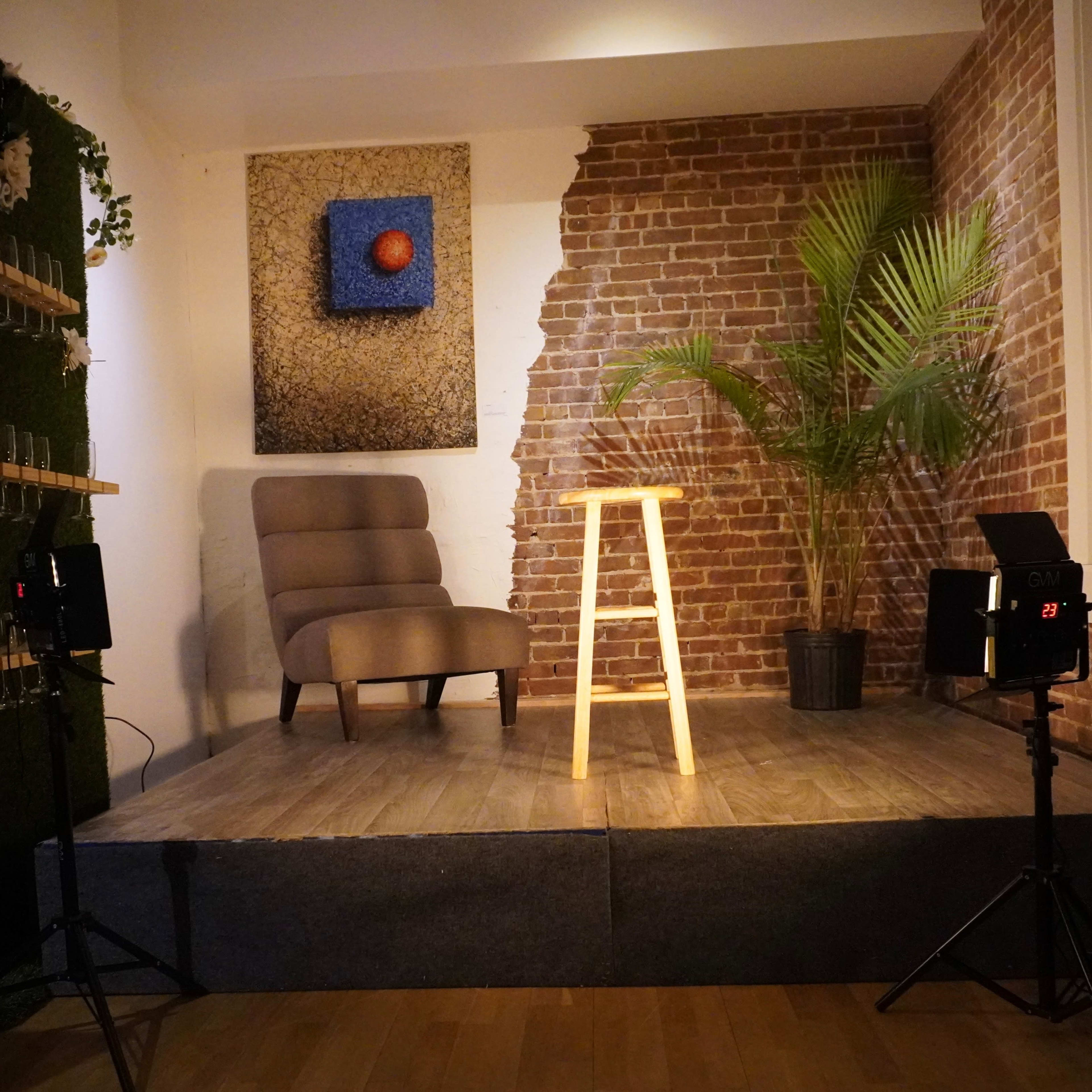 A small stage with a wooden stool, surrounded by a brick wall and a green plant, with lighting equipment positioned nearby.