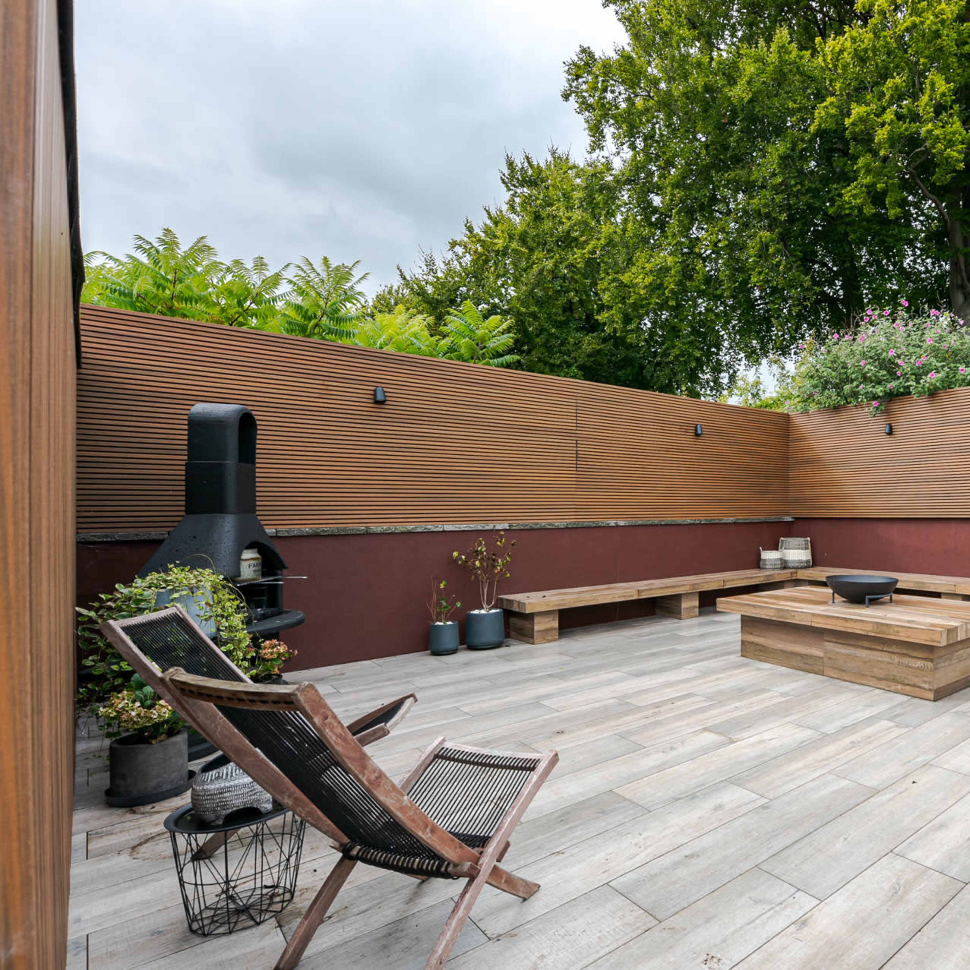 A modern outdoor patio features a wooden seating area with a central table, surrounded by planters and a grill, enclosed by slatted wood walls and greenery.