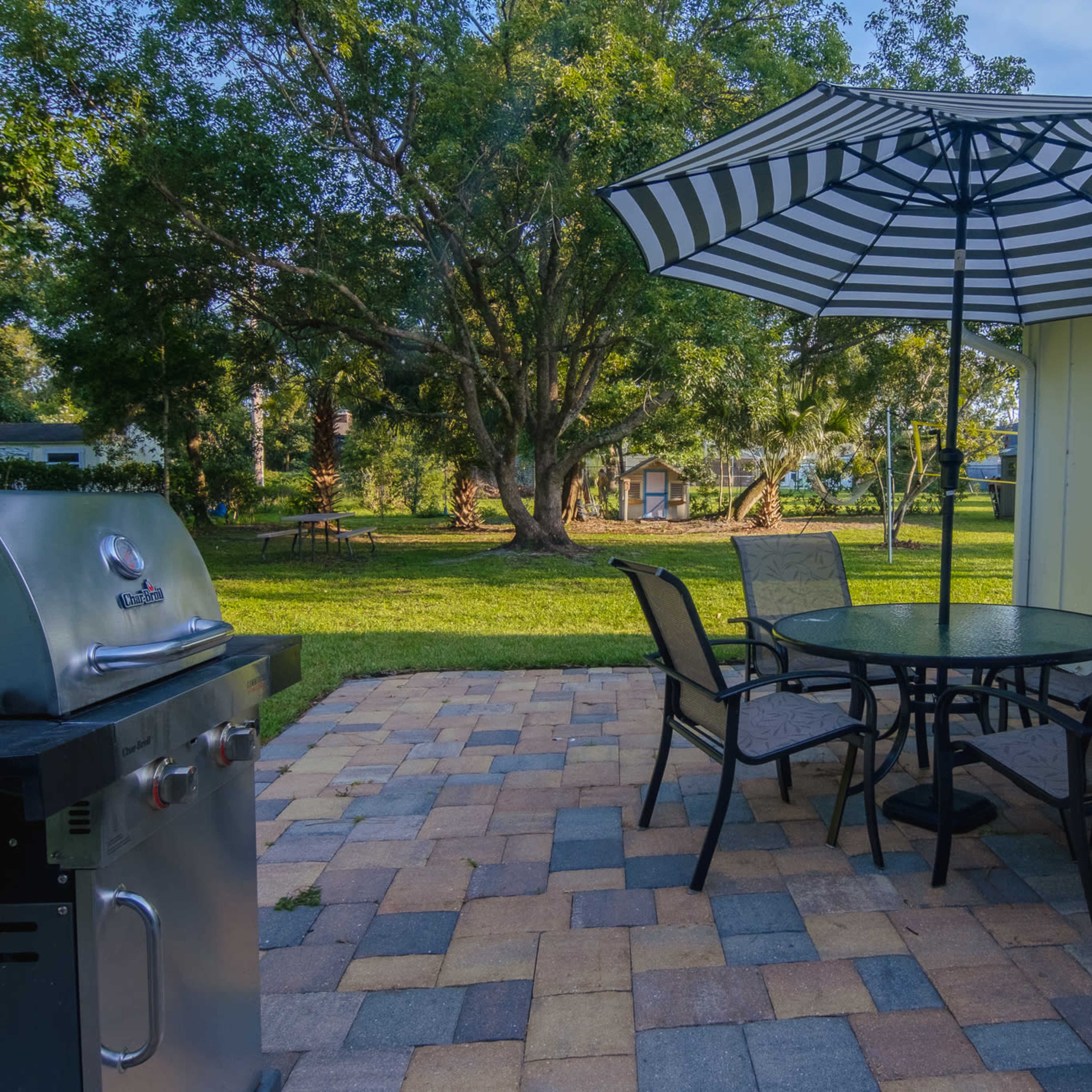 A patio area features a round table with four chairs and a striped umbrella beside a grill, surrounded by a grassy yard and trees.