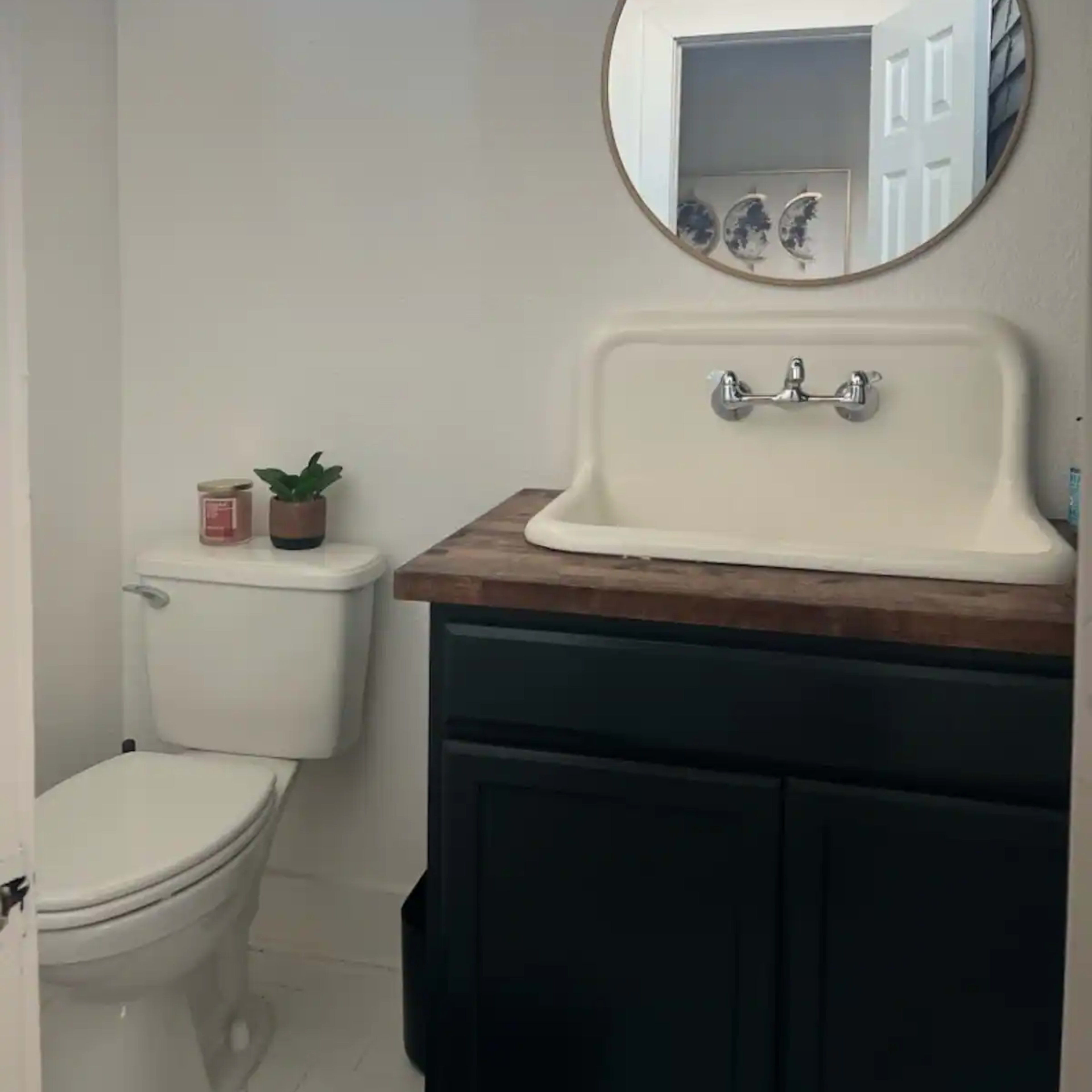 A small bathroom featuring a white sink with a wooden countertop, a toilet, a round mirror, and a plant on the counter.