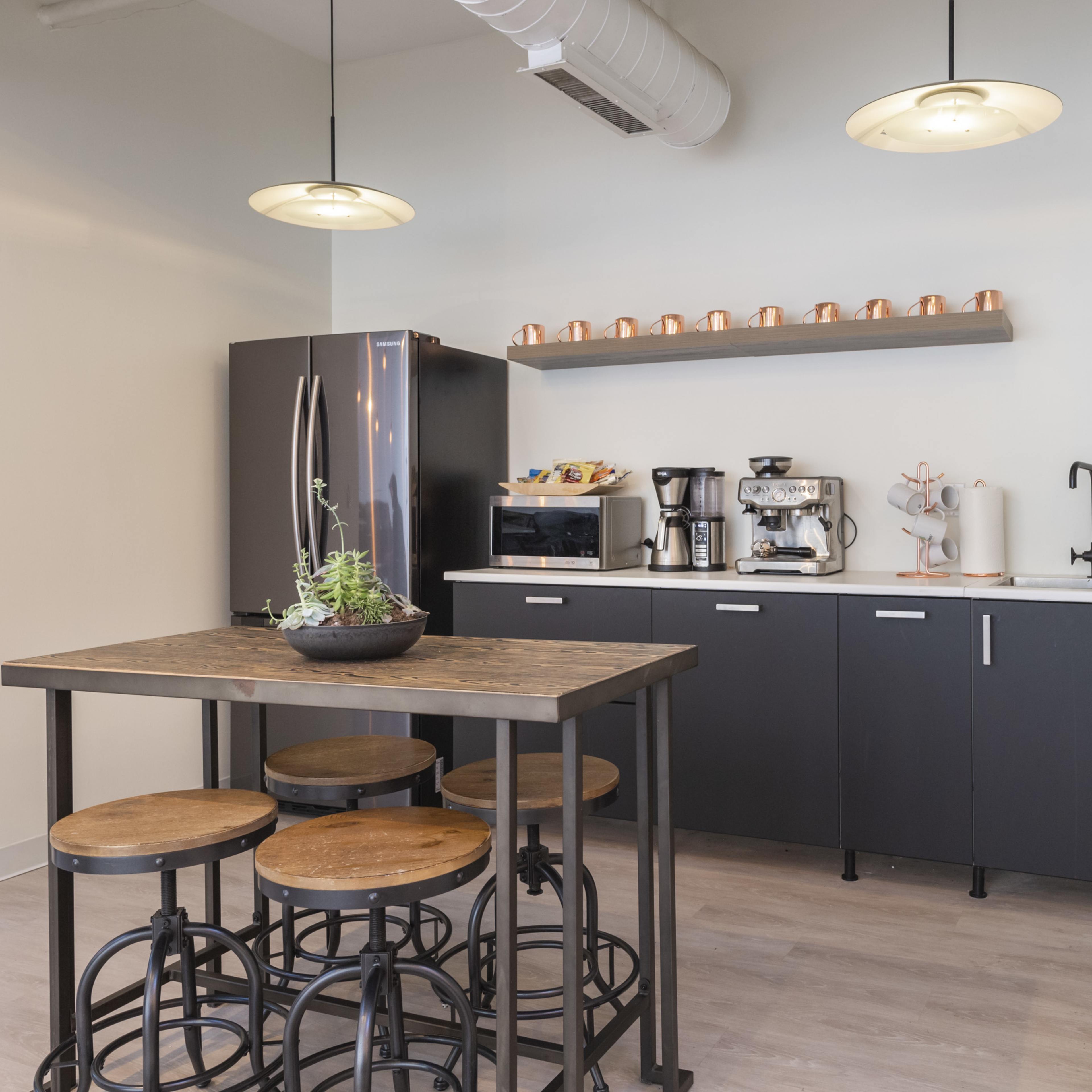 A modern kitchen area with a black refrigerator, coffee machines, and a wooden table surrounded by stools.