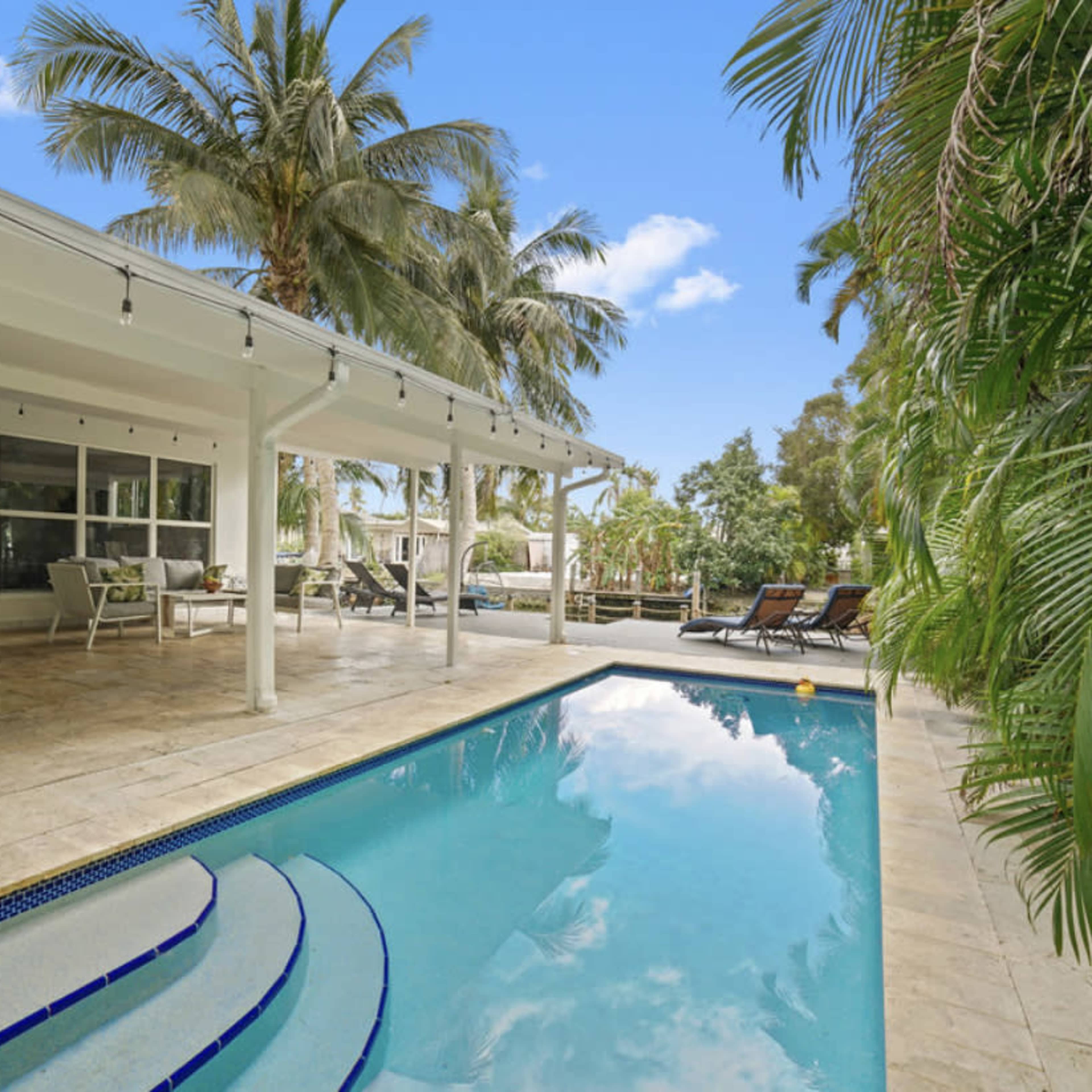 A swimming pool surrounded by a stone patio, with palm trees and lounge chairs in a sunny outdoor setting.