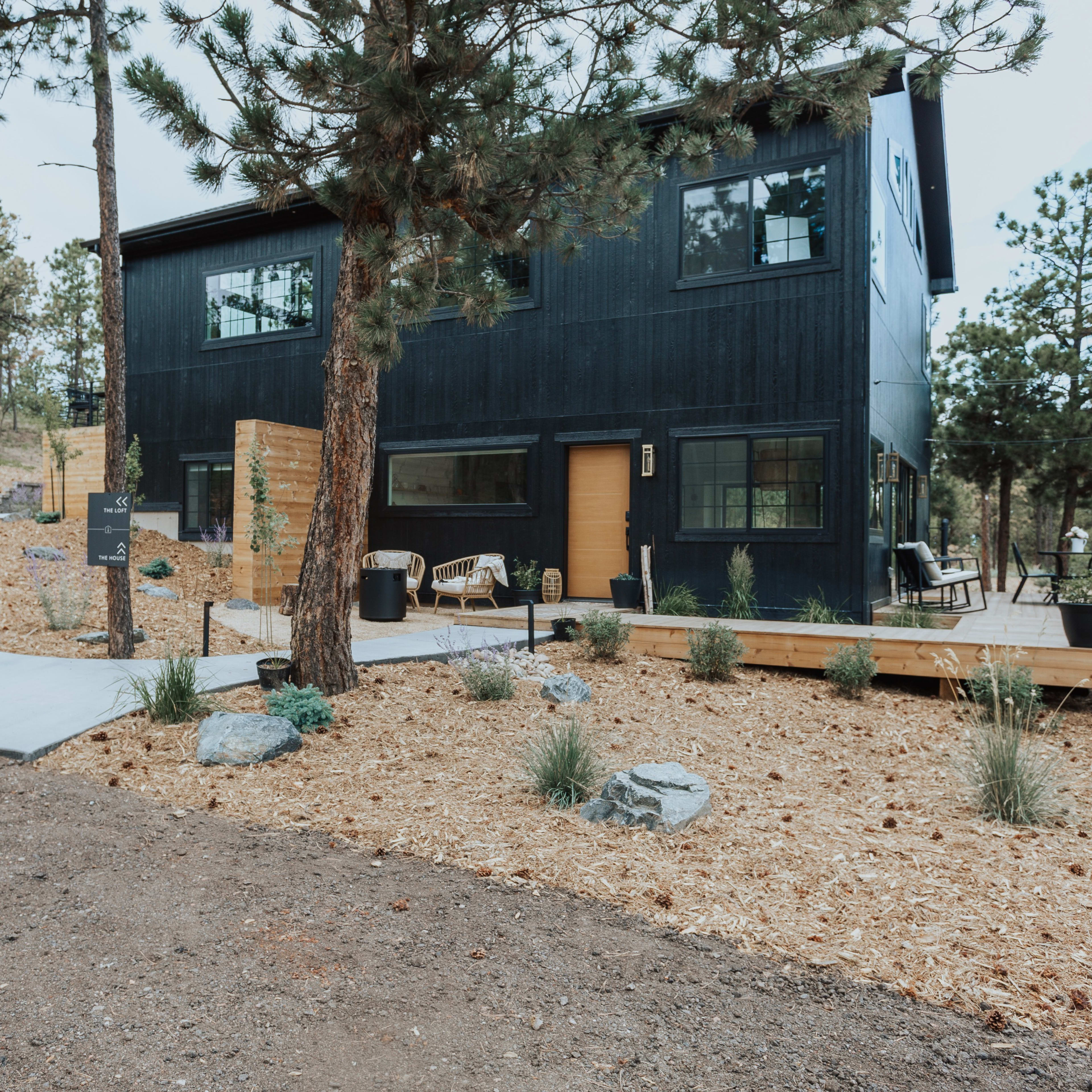 A modern black house with large windows stands surrounded by pine trees and landscaping featuring gravel paths and shrubs.