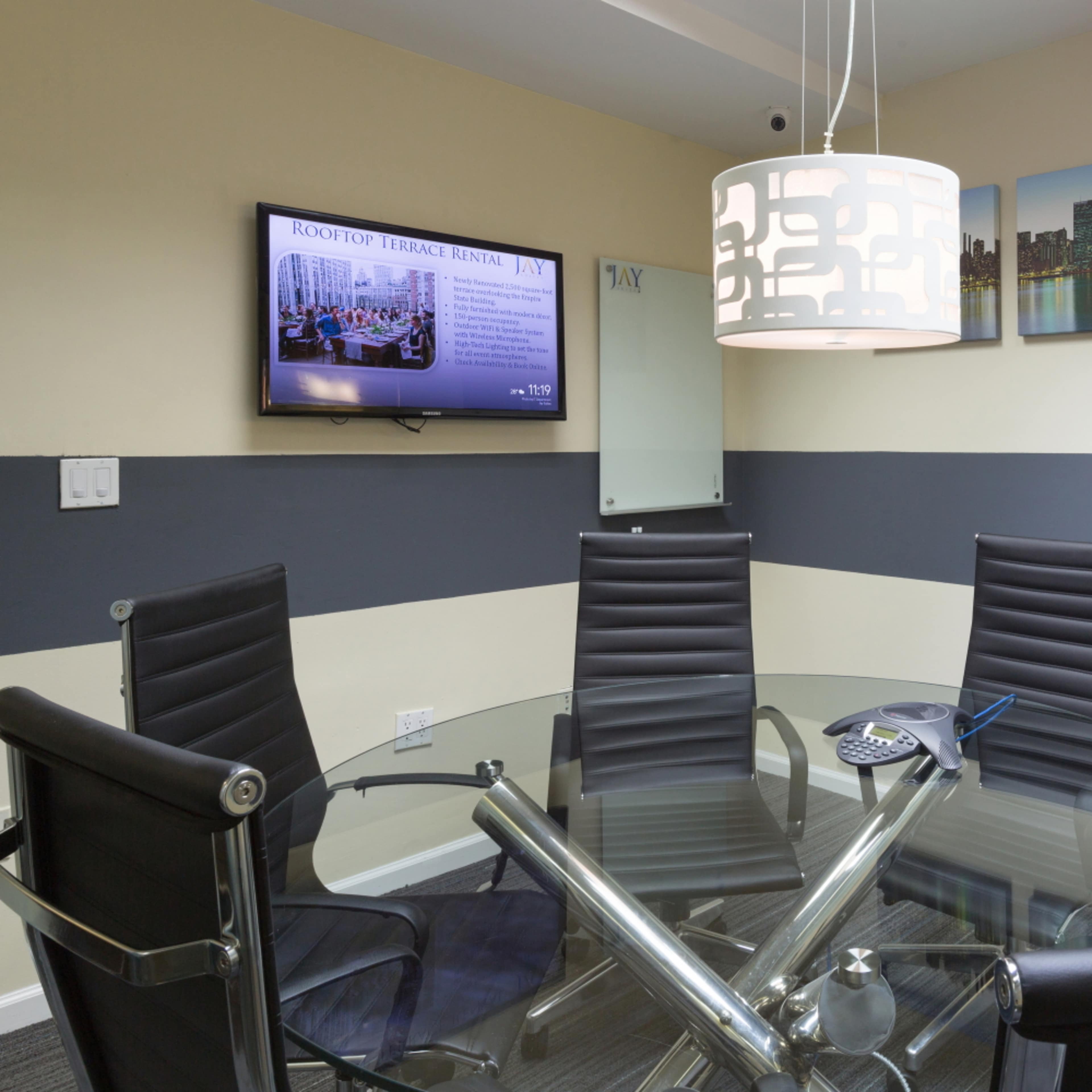 A modern conference room with a glass table, black leather chairs, a television mounted on the wall, and cityscape artwork.