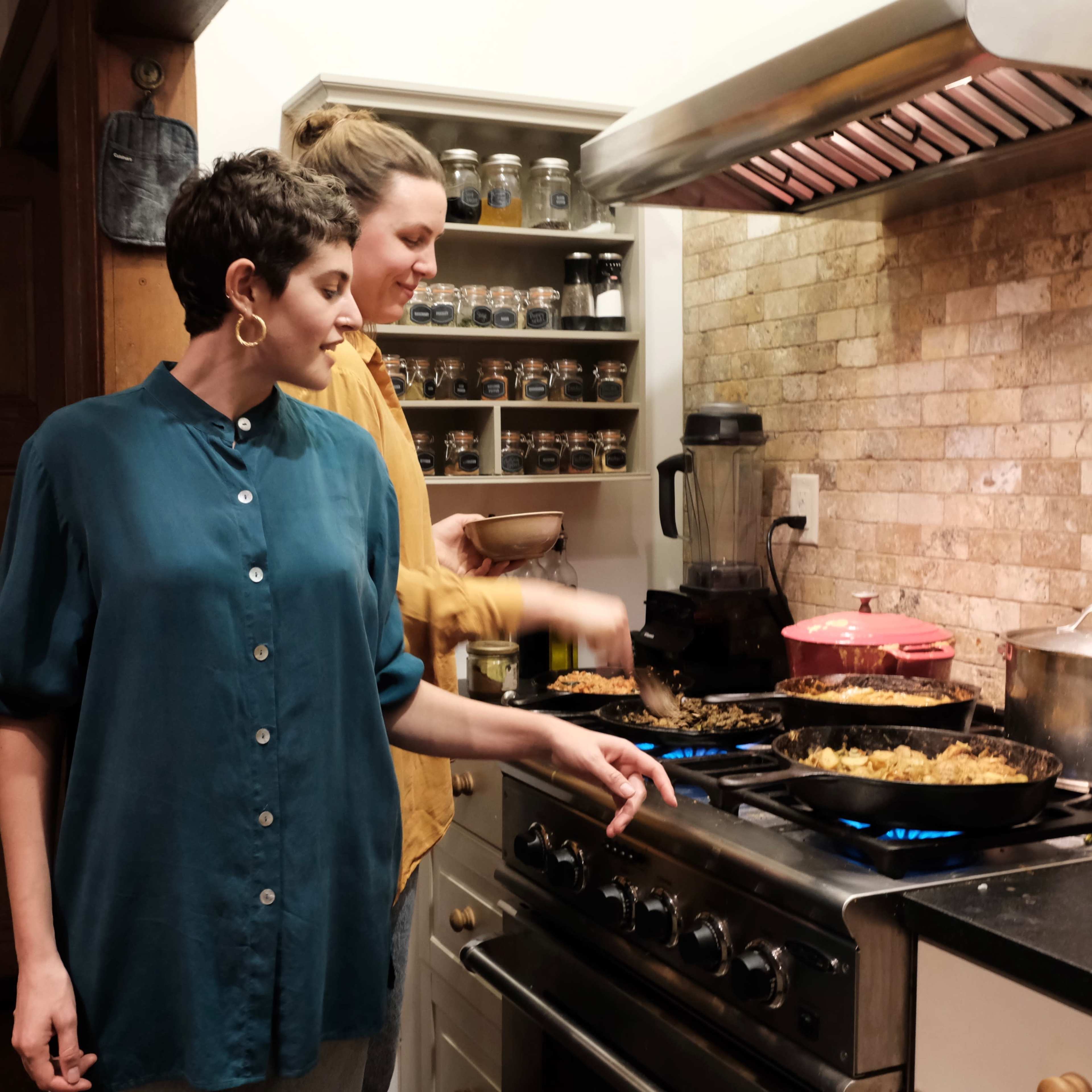 Two women prepare food in a kitchen, using multiple frying pans on the stove.