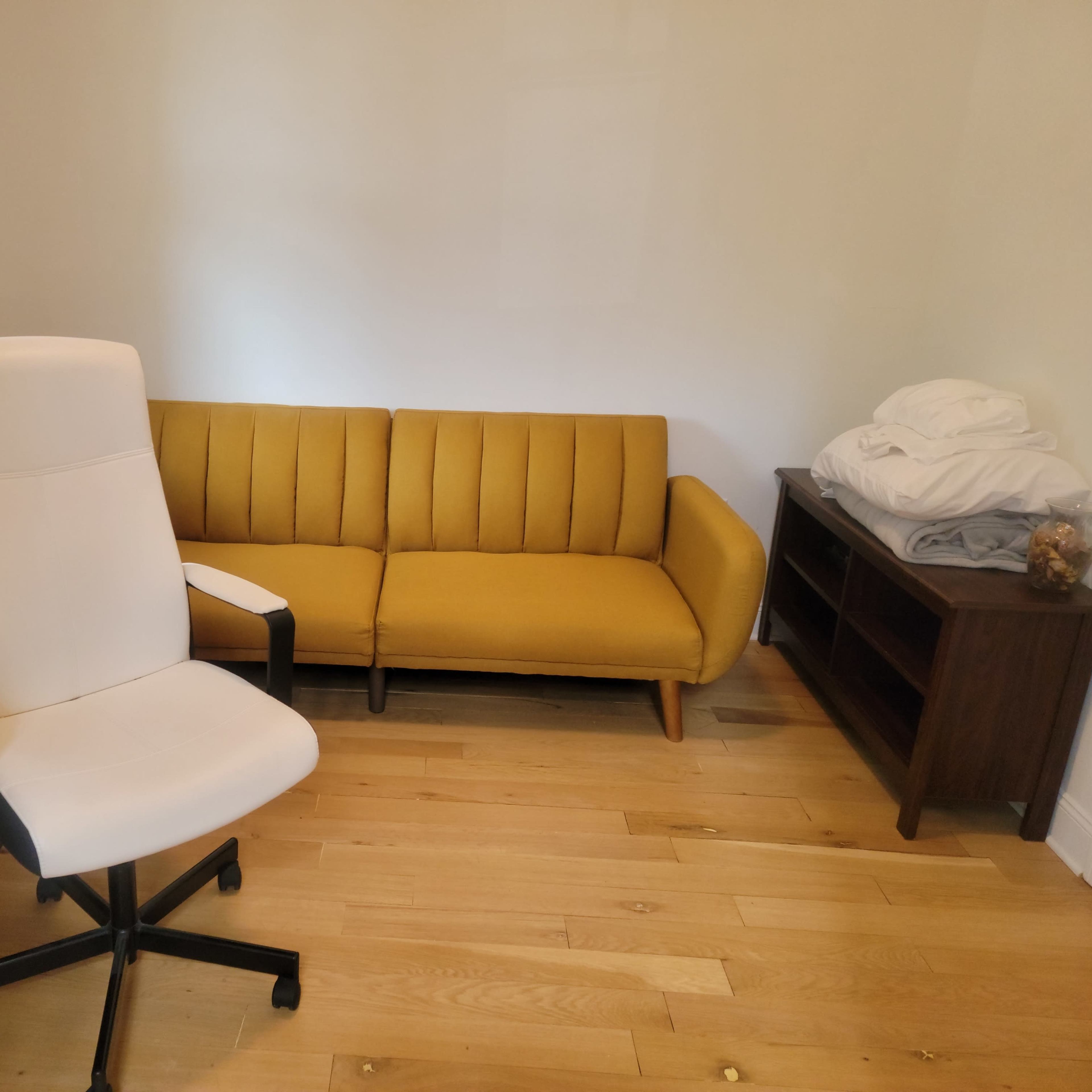 The room features a yellow couch, a white ergonomic chair, a wooden desk, and a cabinet with a few items inside.