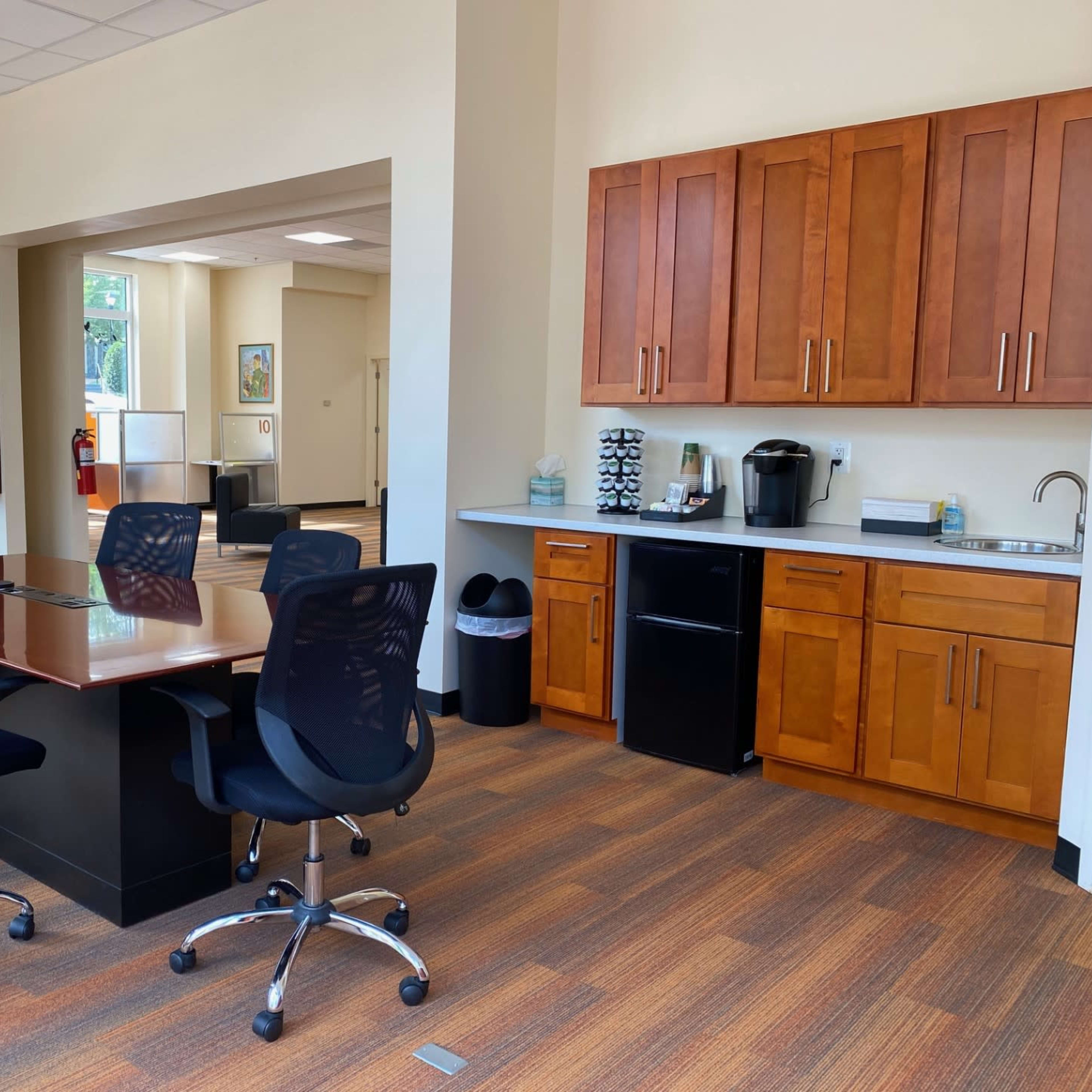 A conference room with a dark wood table surrounded by black office chairs, adjacent to a kitchenette featuring wooden cabinets and appliances.