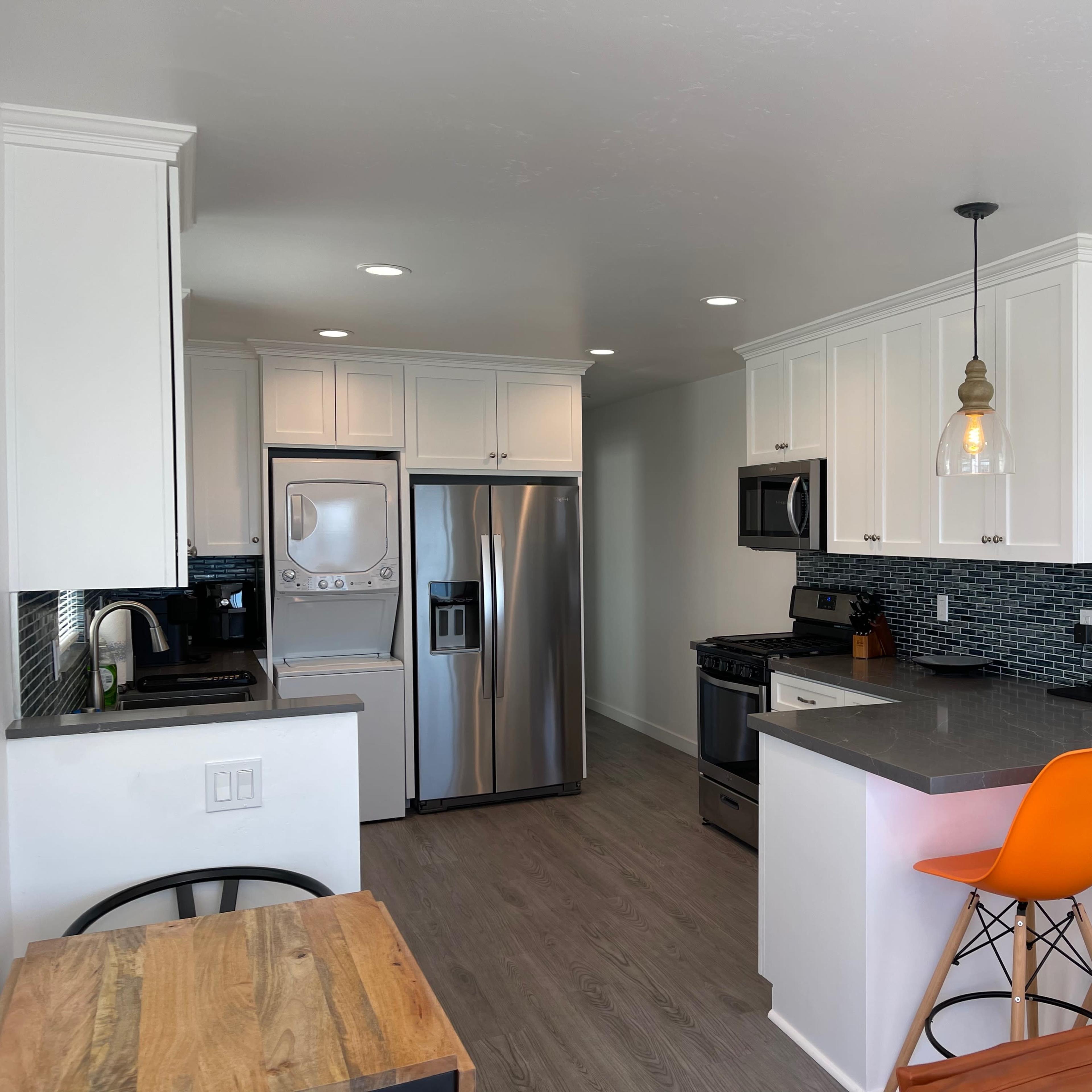 A modern kitchen with stainless steel appliances, white cabinetry, and a small dining area featuring wooden and orange bar stools.