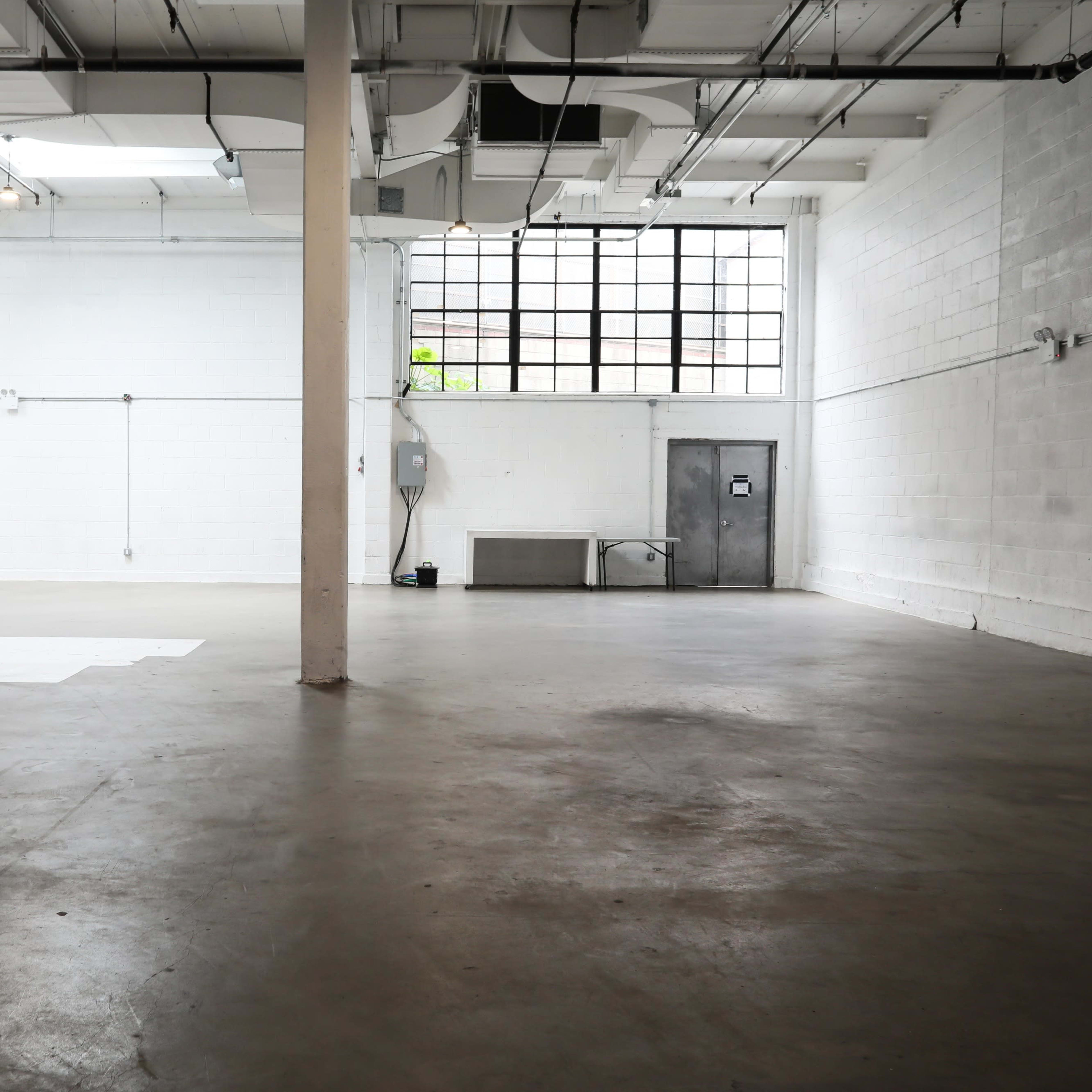 A spacious, empty interior of a warehouse or studio with concrete flooring and a large window on one wall.