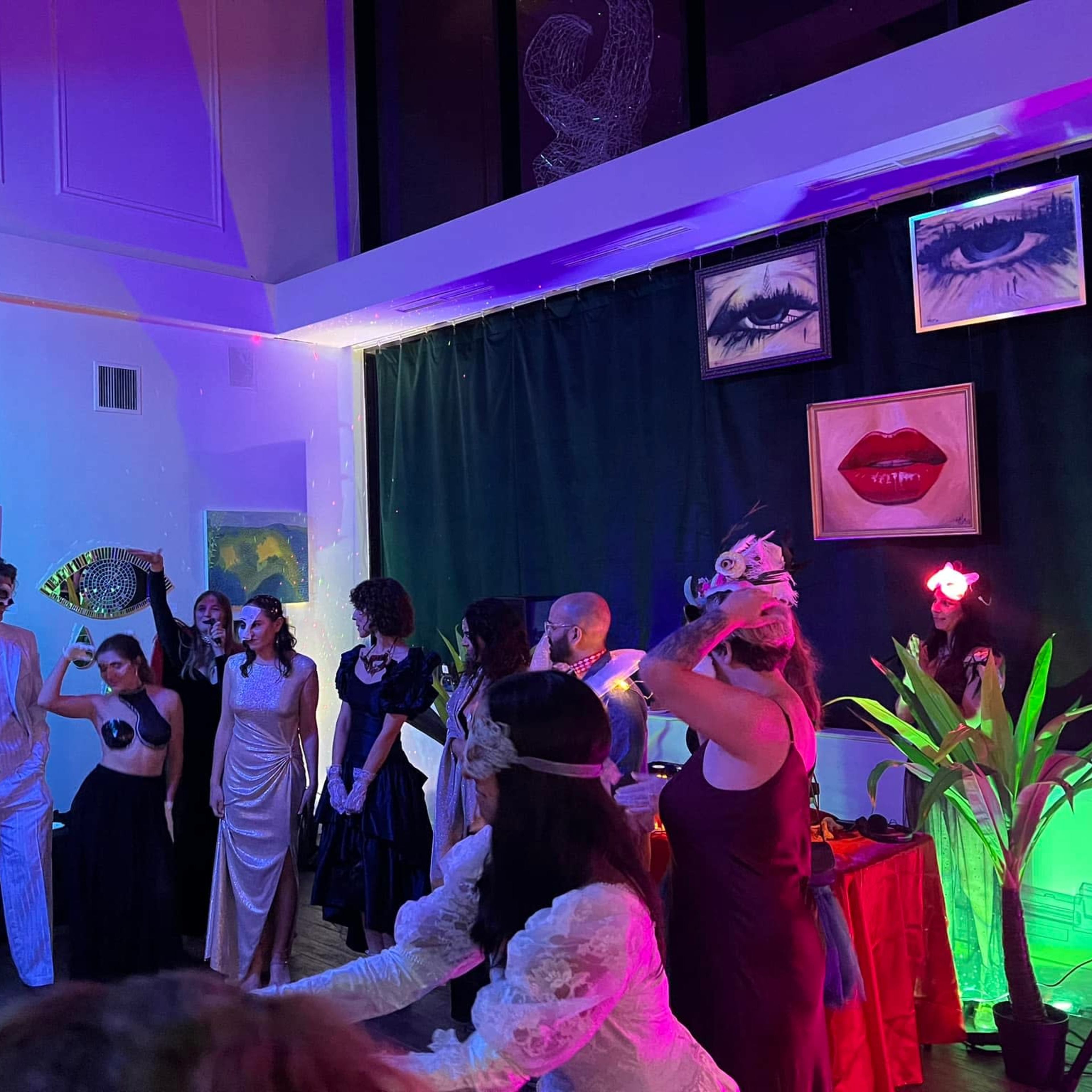 A group of elegantly dressed guests gathers in a brightly lit room adorned with vibrant colors and framed portraits, preparing for a themed event.