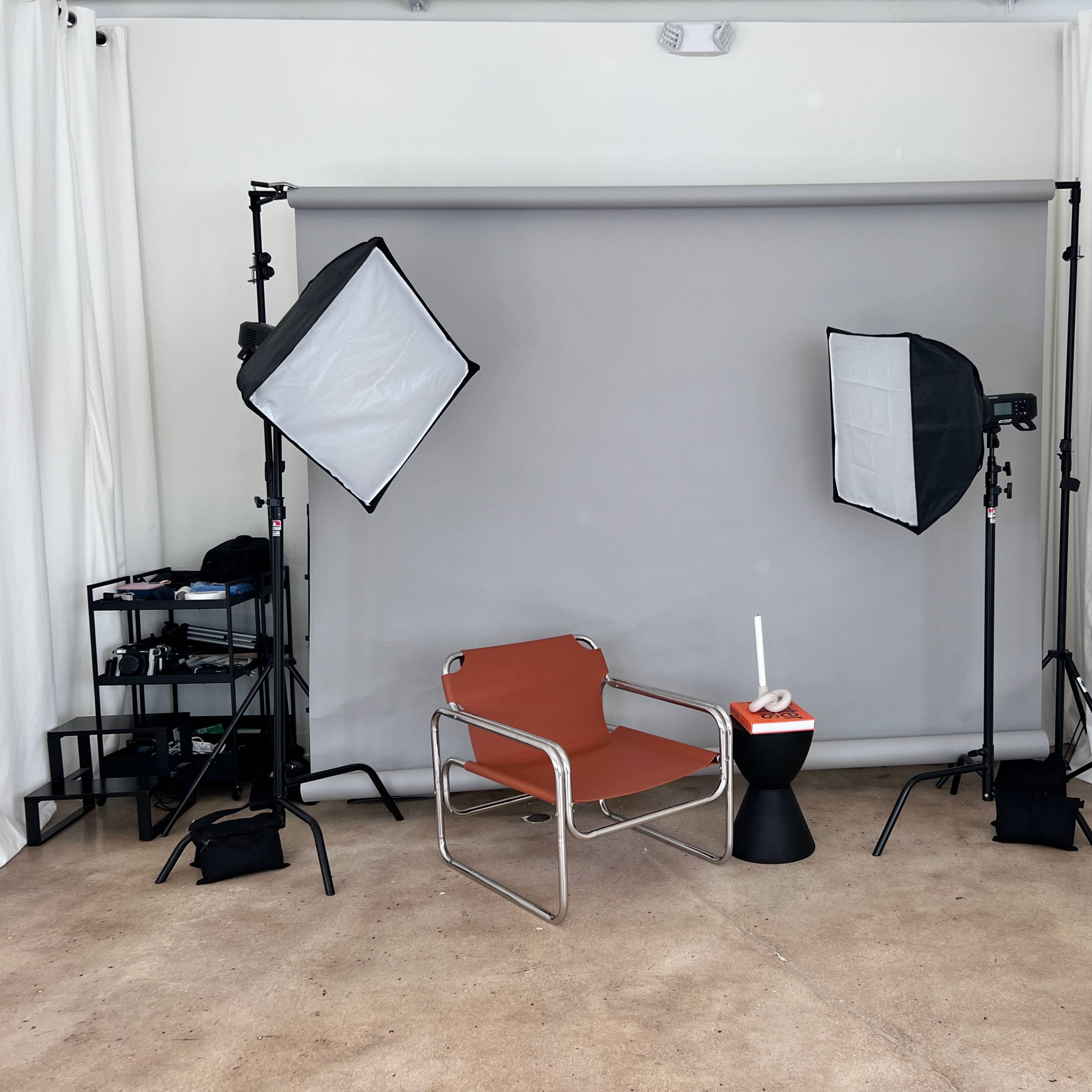 A minimalist photography studio setup with gray backdrop, two softbox lights, a chair, and a small black table in the center.