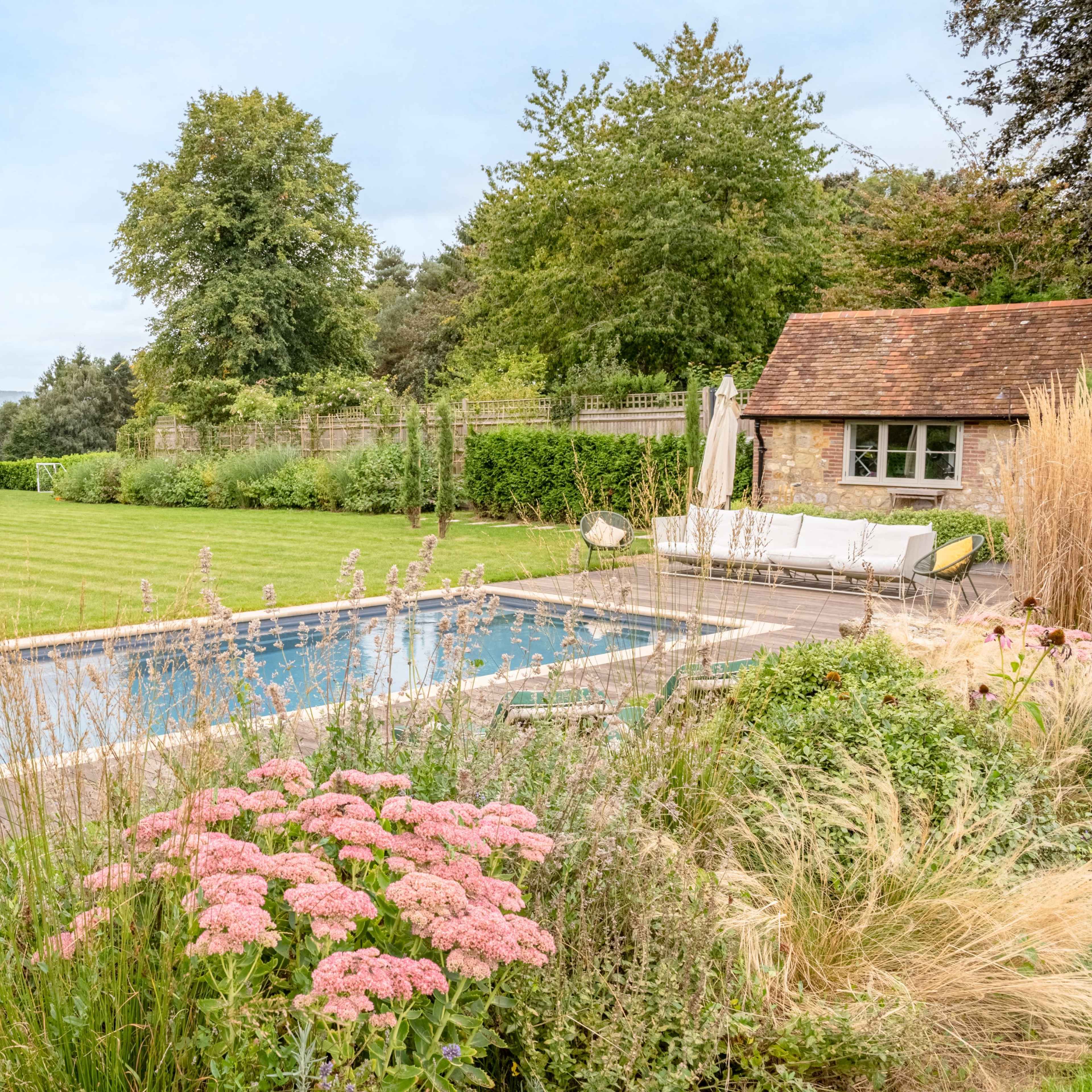A rectangular swimming pool with lounge chairs is surrounded by neatly trimmed grass and flower beds in a landscaped garden.