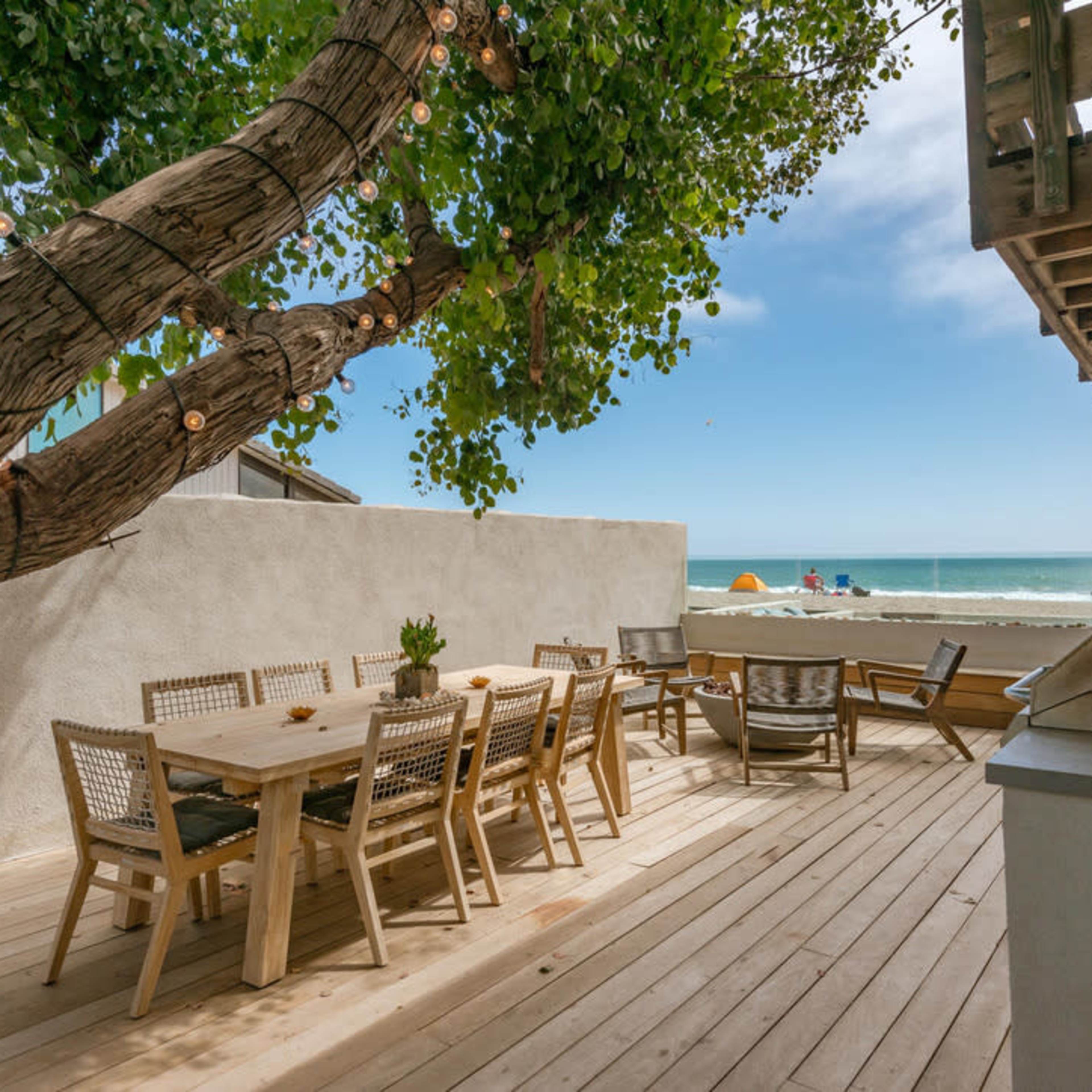 A wooden deck with a dining table and chairs overlooks the ocean, with a tree providing shade and string lights hanging above.