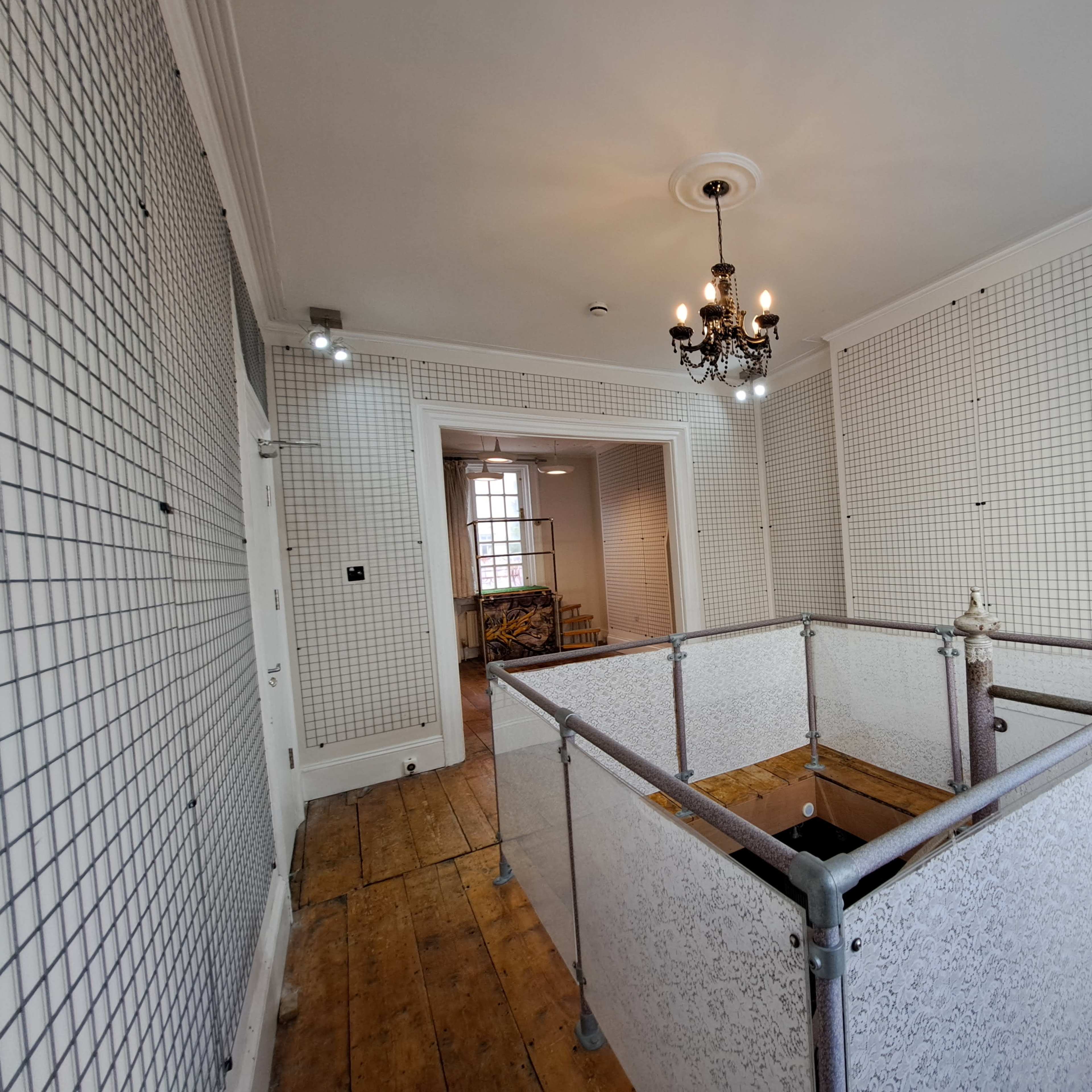 A spacious room with wooden flooring, grid-patterned wall panels, and a decorative chandelier, featuring a partial enclosure made of white mesh and metal.