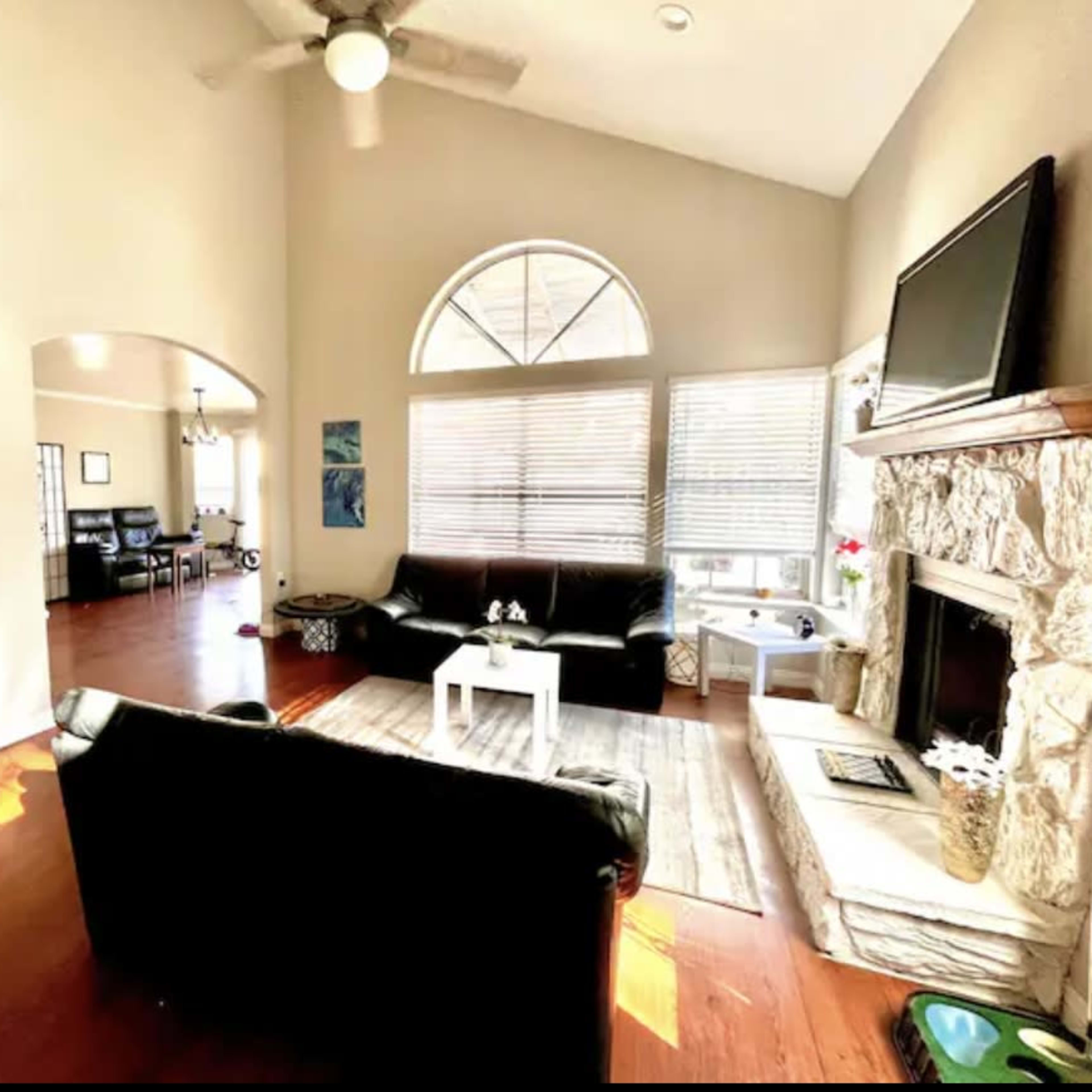 A spacious living room featuring a black leather sofa, a stone fireplace, and large windows that let in natural light.