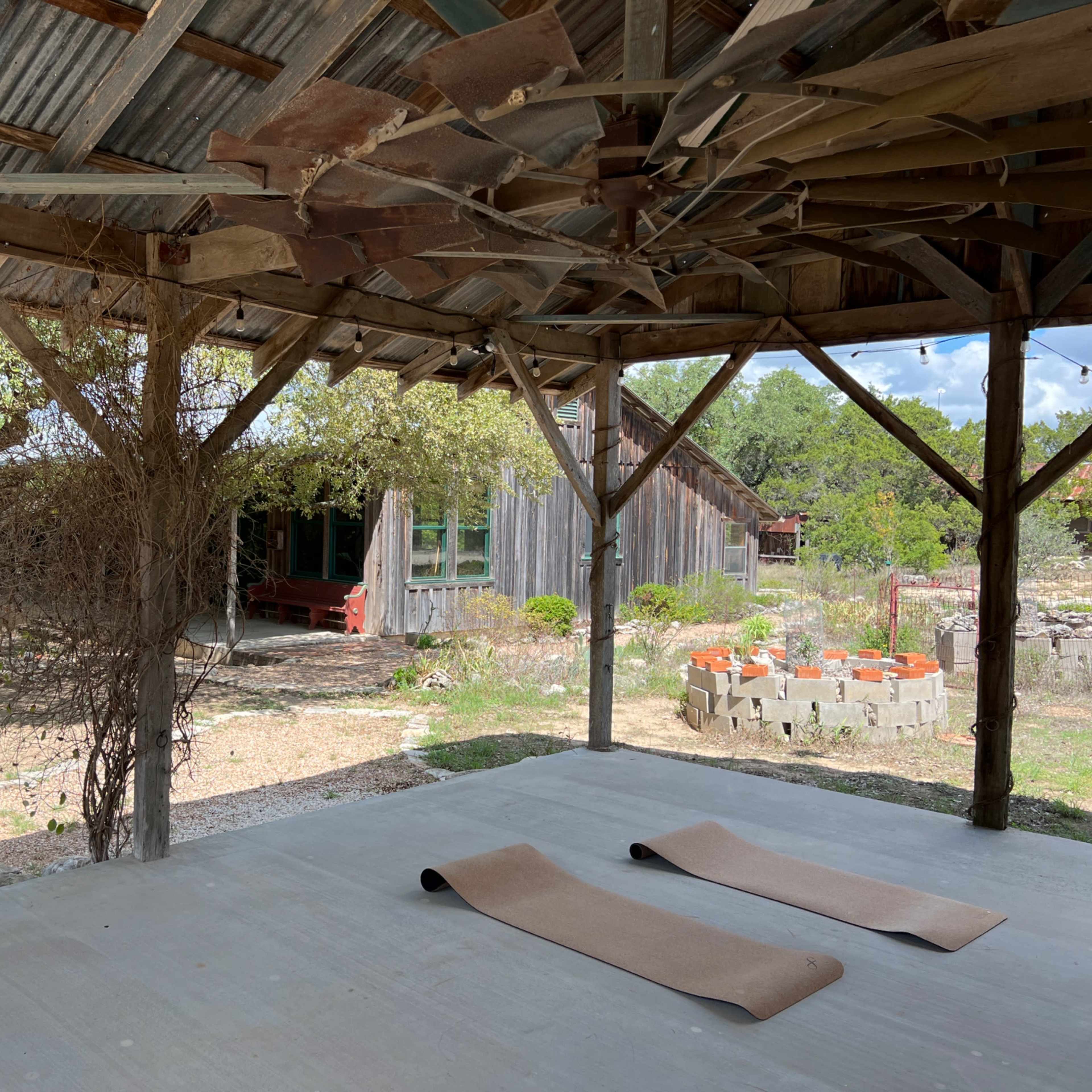 An outdoor space with two yoga mats on a concrete floor under a wooden roof, overlooking a garden area with rustic structures in the background.