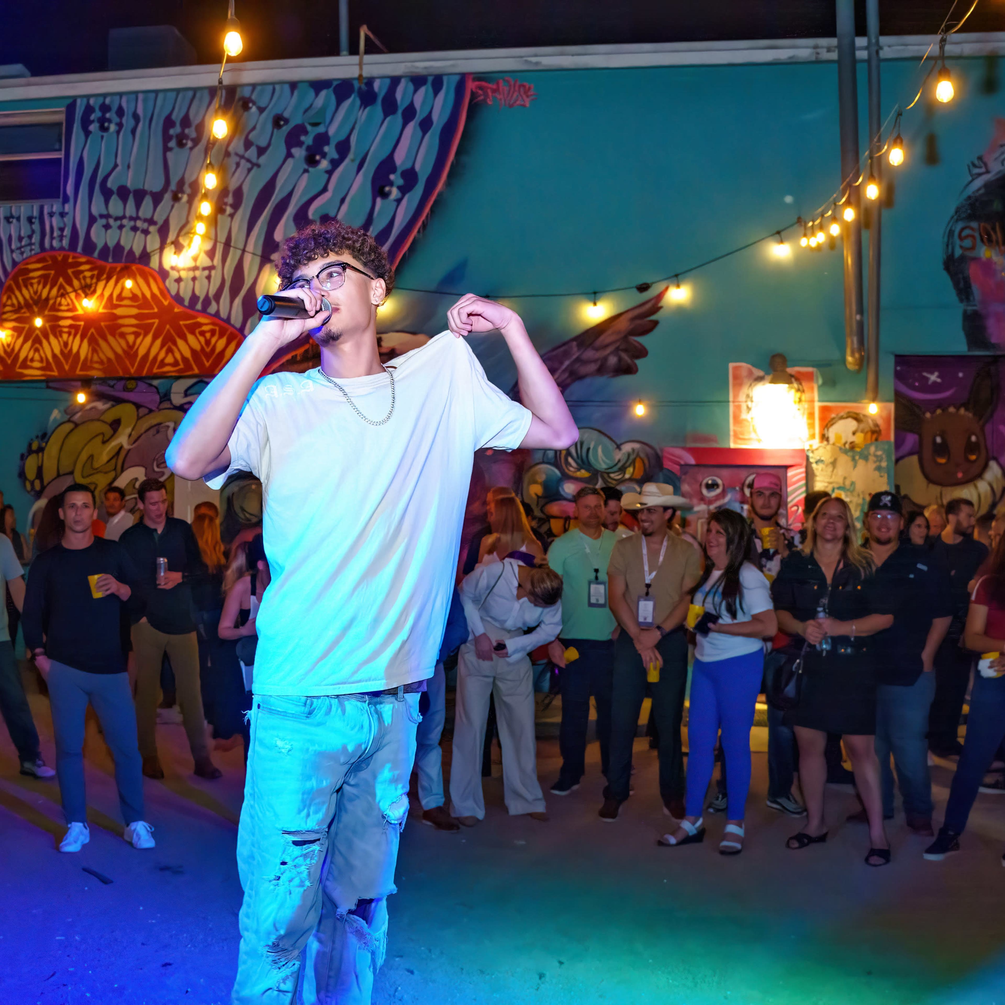 A young man performs with a microphone in front of an audience in an outdoor space adorned with colorful murals and string lights.