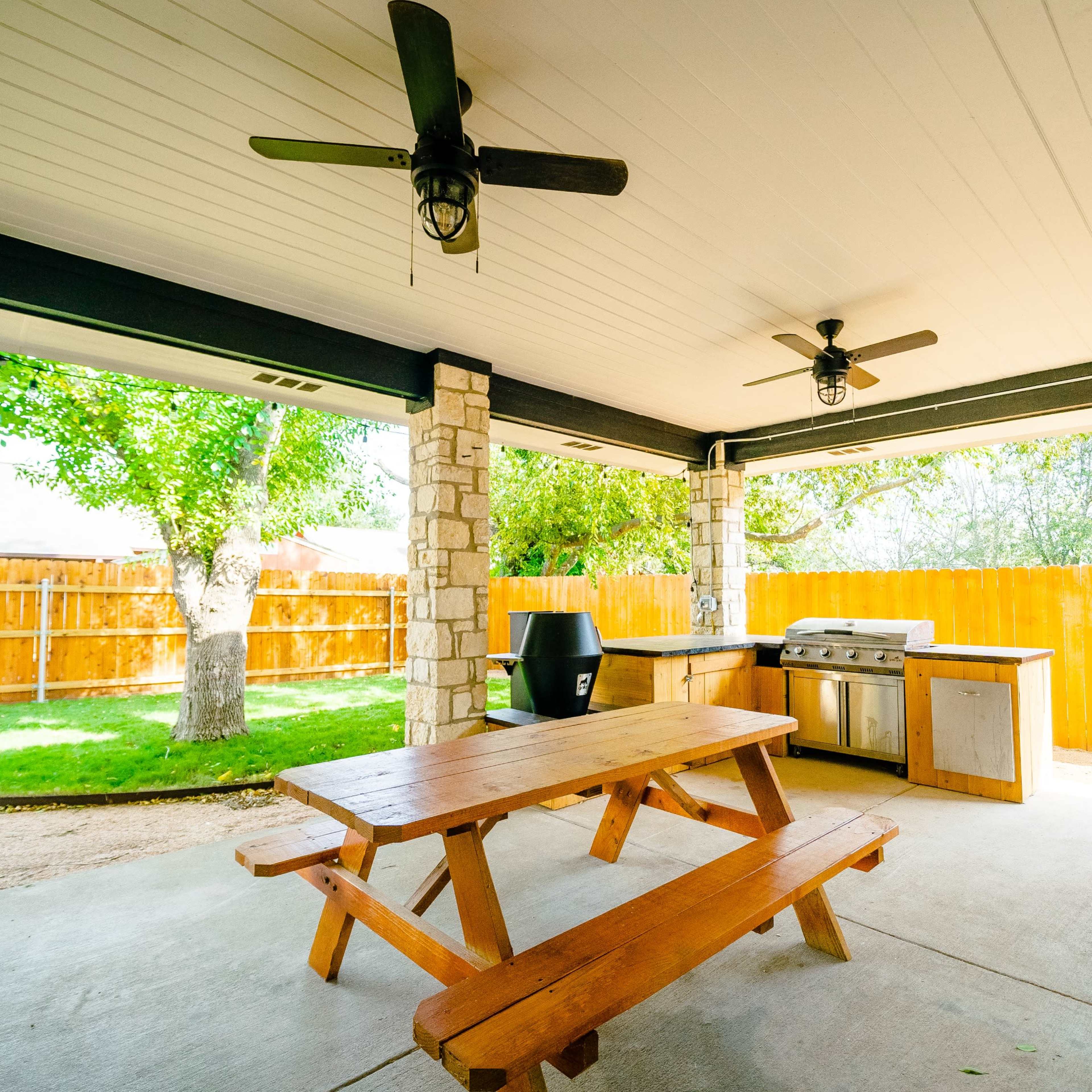 A covered outdoor patio with a wooden picnic table, a barbecue grill, and two ceiling fans, surrounded by a fenced yard with greenery.