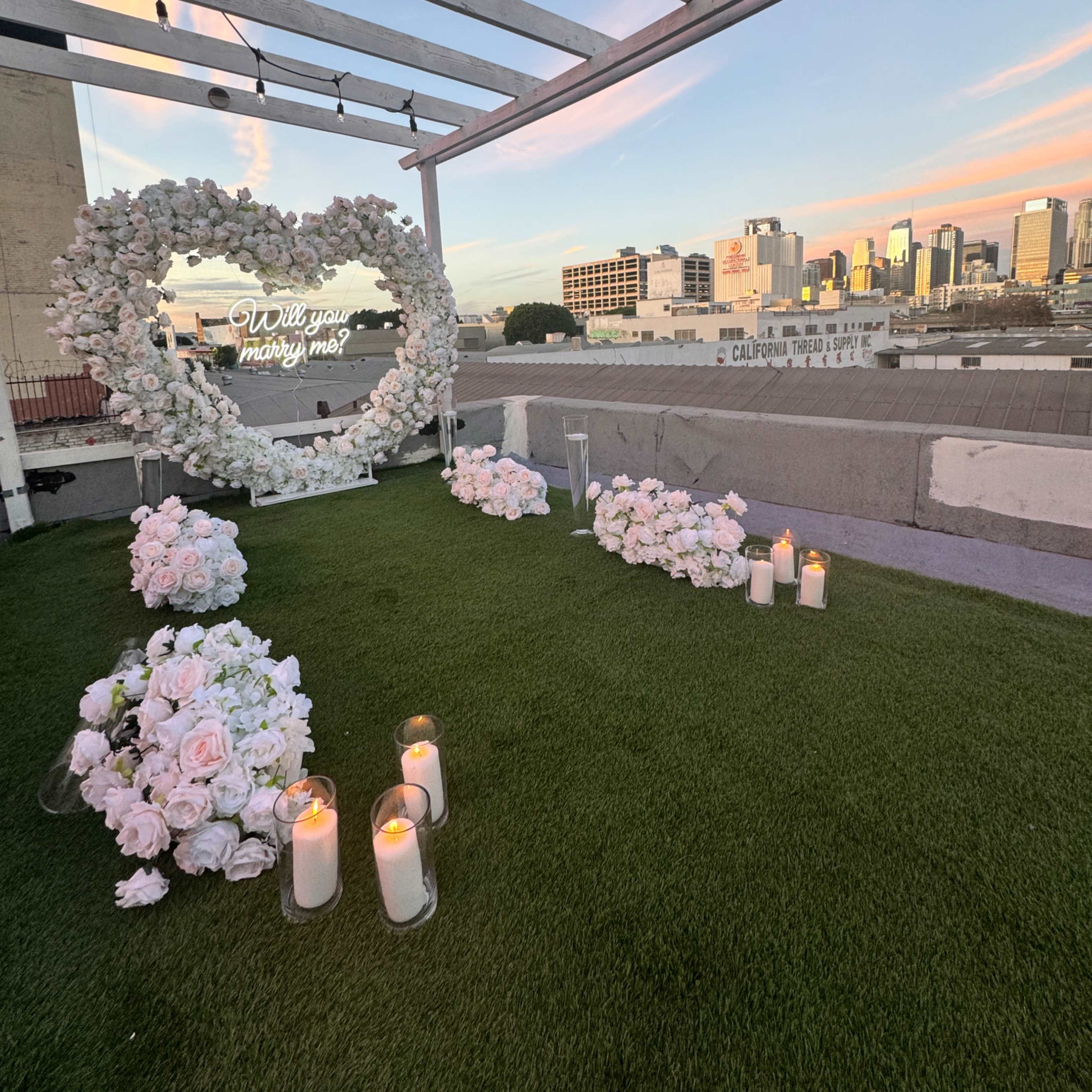A heart-shaped floral arrangement with candles and a sign reading "Will you marry me?" is set on a rooftop with a skyline view at sunset.