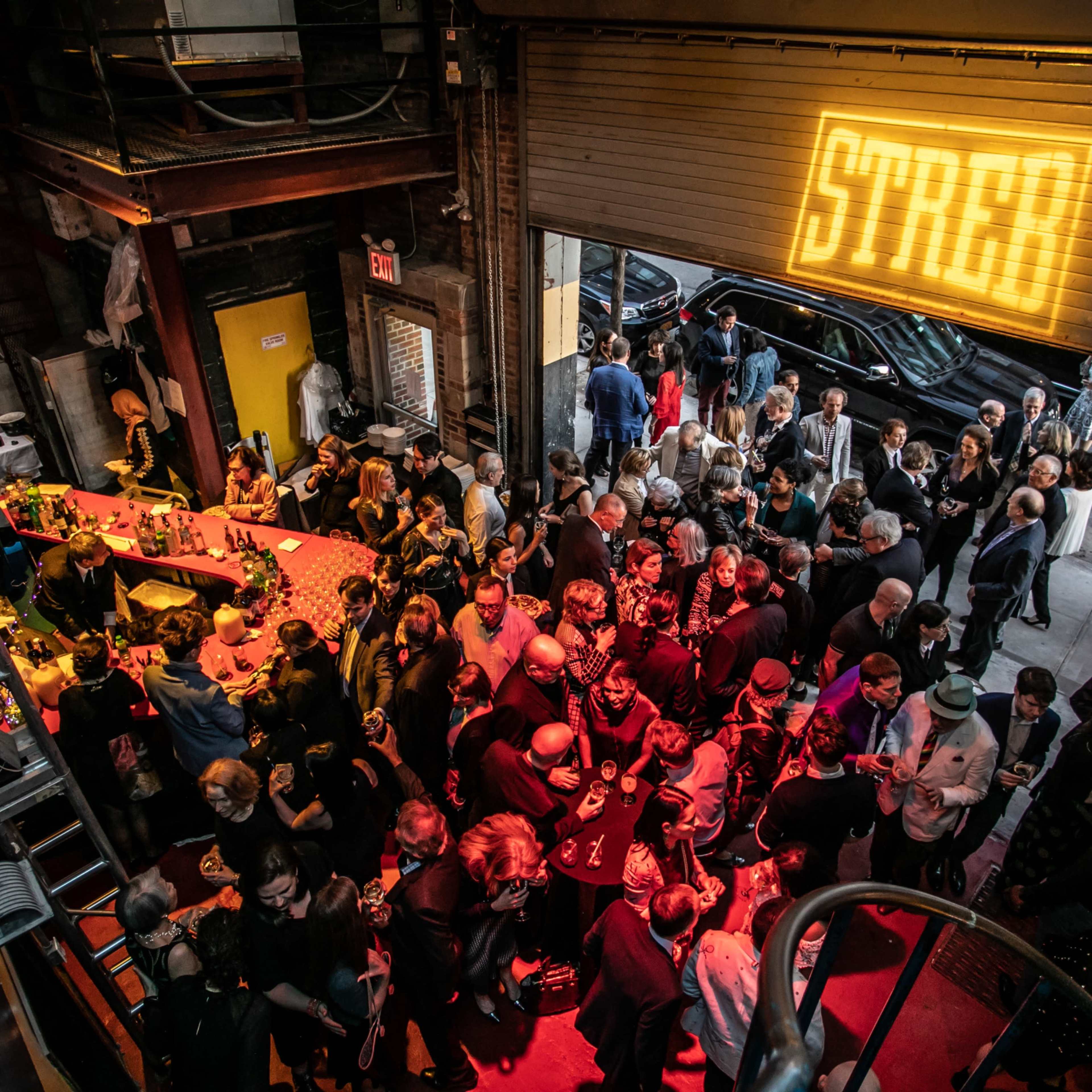 A large crowd gathers inside a venue with a bar, illuminated by a sign that reads "STREET," while a car is parked outside.