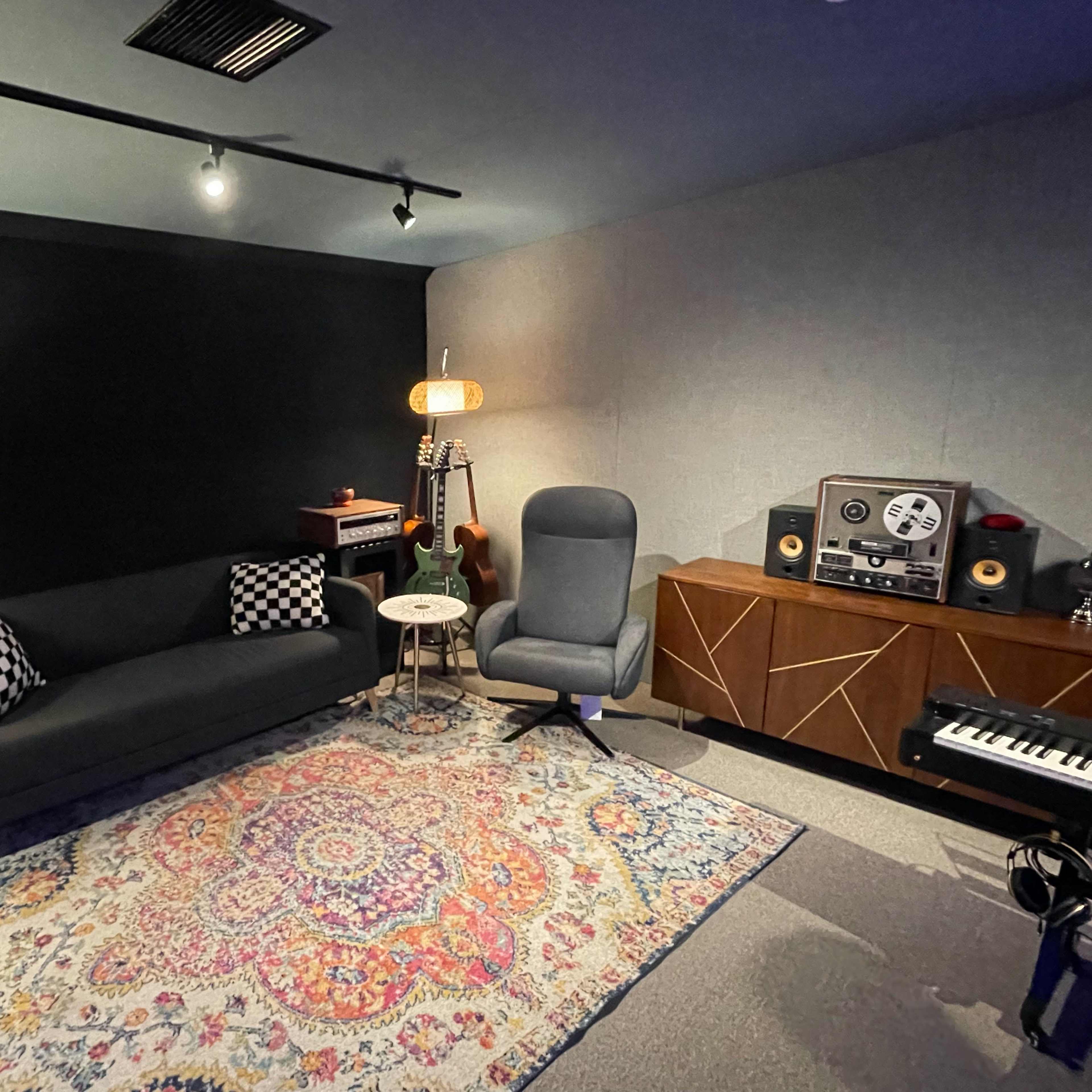 A cozy studio room featuring a black sofa, a patterned rug, a small table, a lamp, musical instruments, and audio equipment.