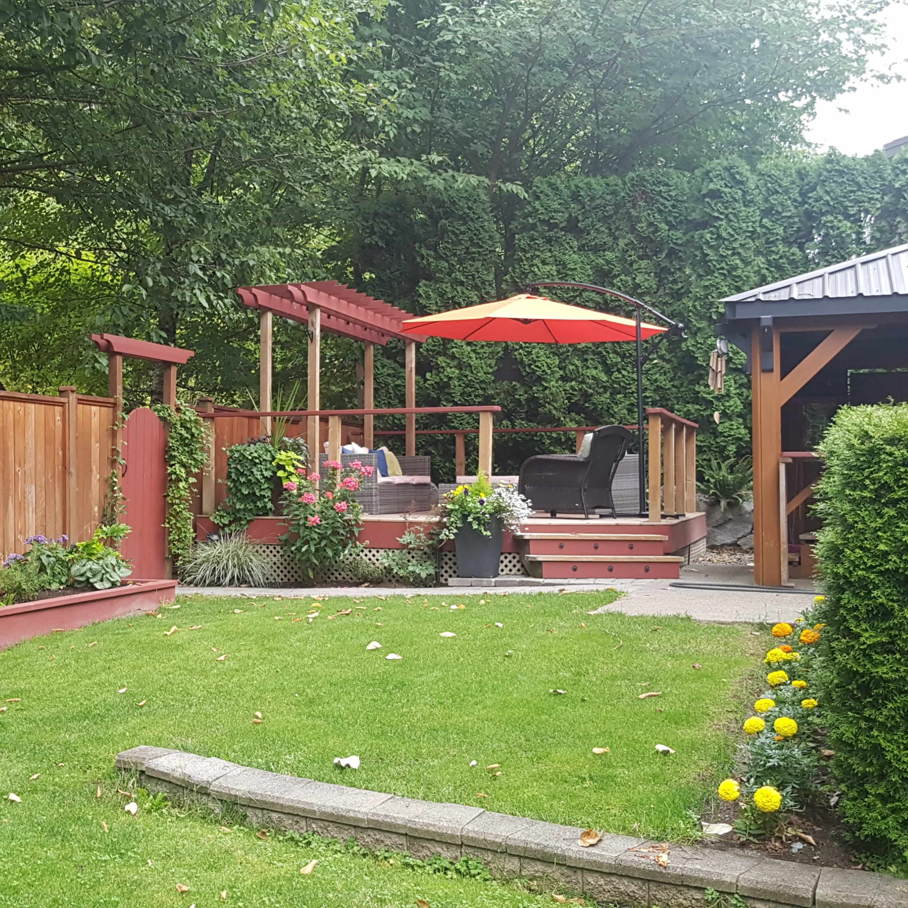 A landscaped backyard with wooden fencing, a deck featuring outdoor seating under an orange umbrella, and flower beds flanking a grassy area.