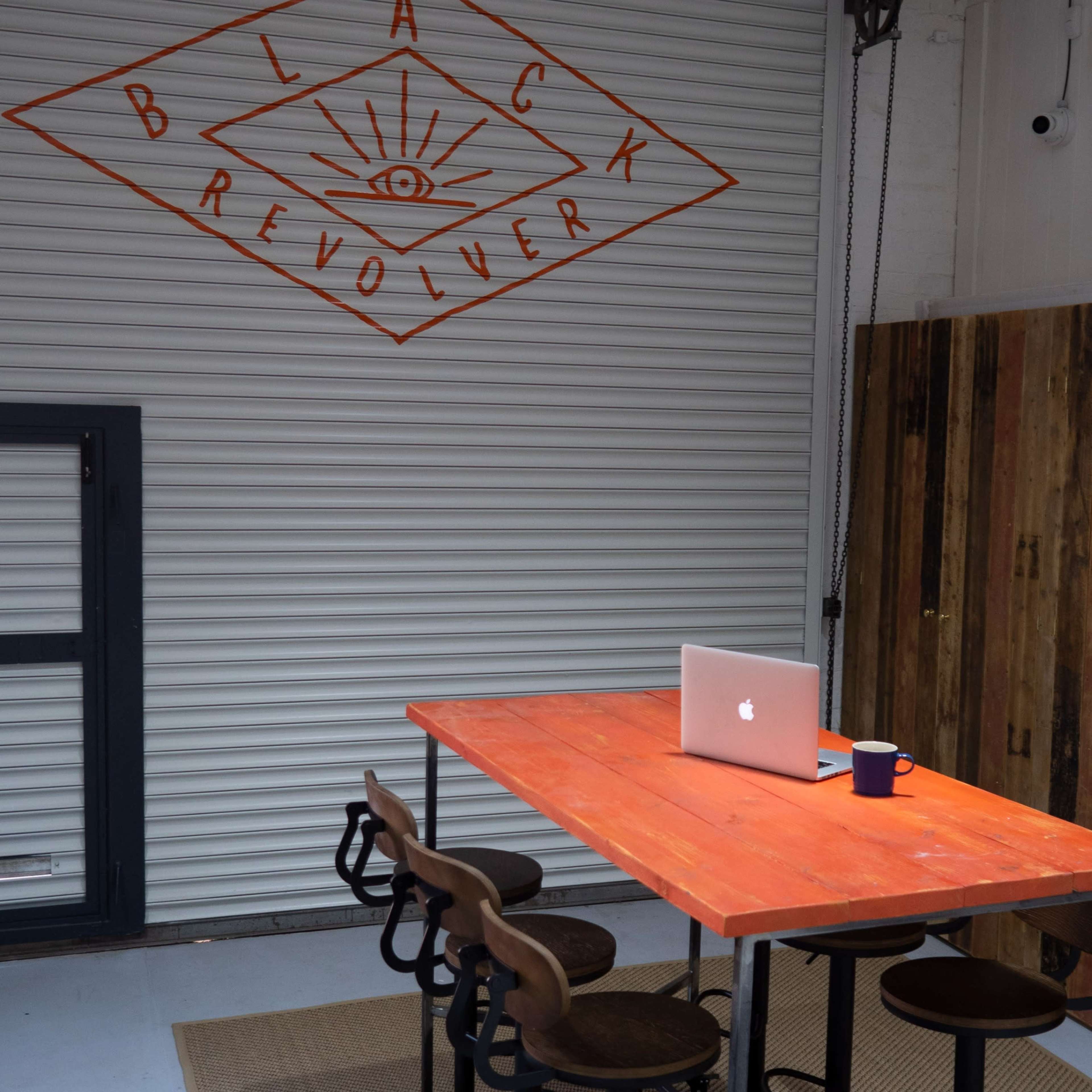 A simple workspace features a vibrant orange table with four black stools, a laptop, and a coffee mug, against a backdrop of a closed garage door with an orange graphic design.