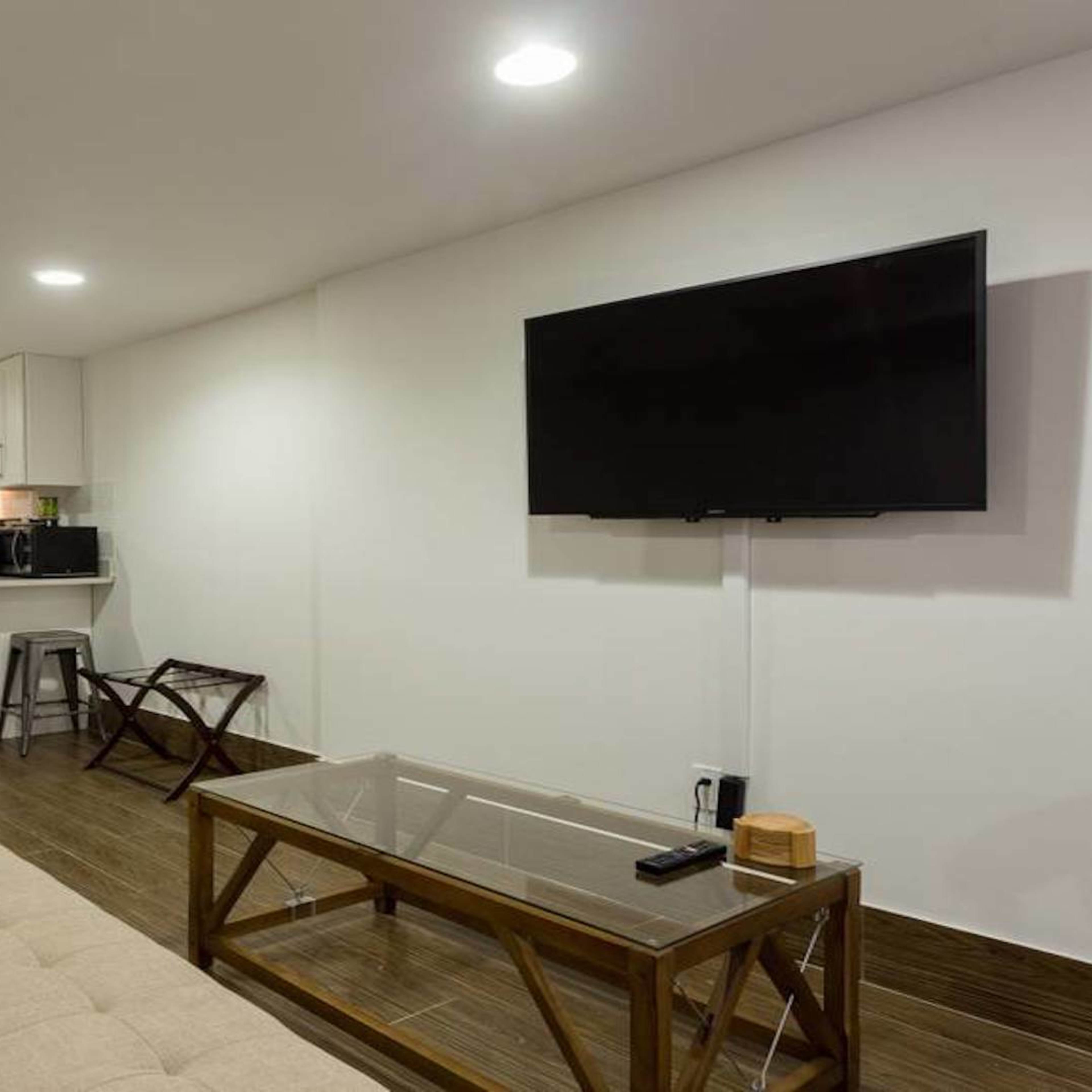A modern living room with a beige sofa, a glass coffee table, a mounted television, and a compact kitchen in the background.