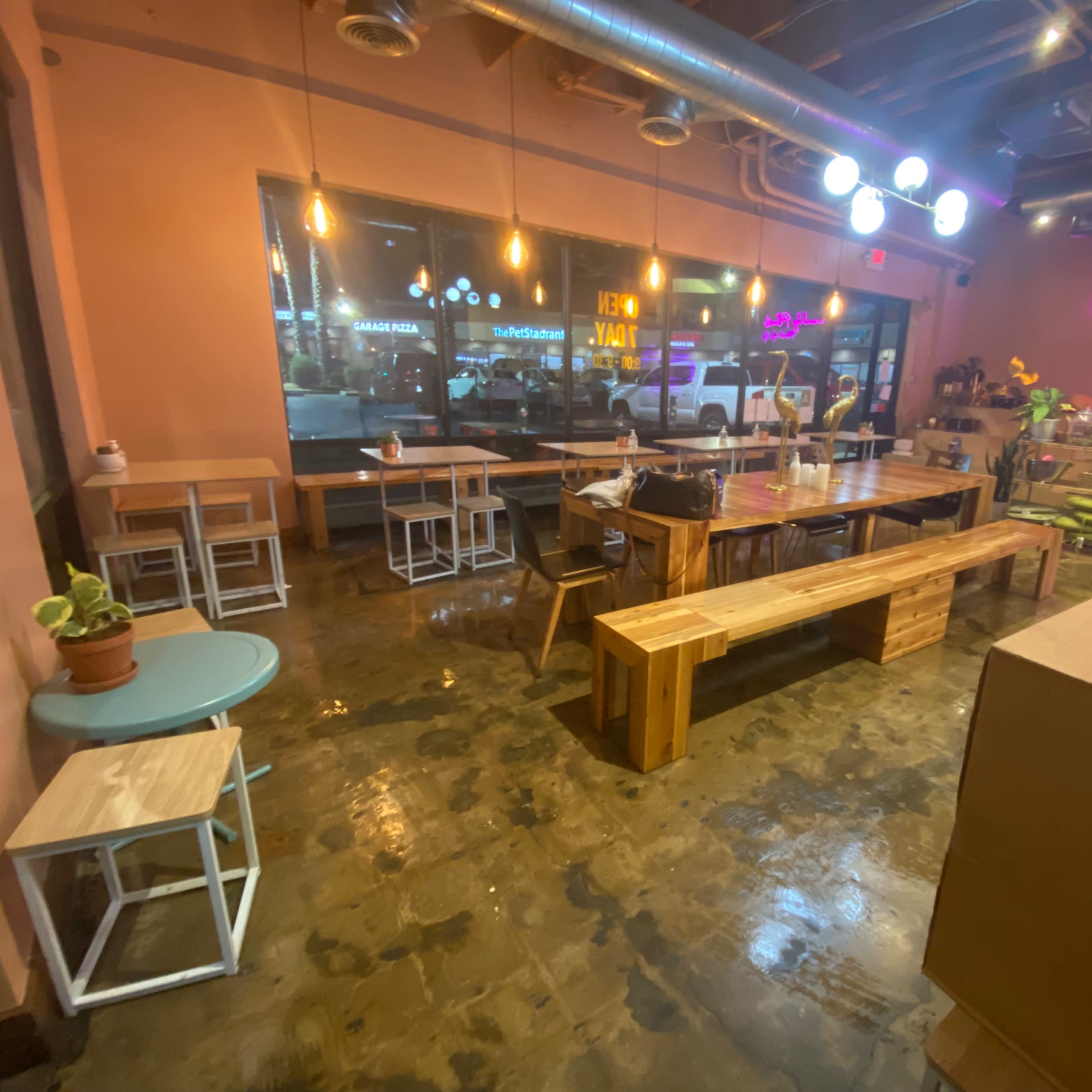 A contemporary café interior with wooden tables and benches, dim lighting, and large windows displaying neon signs outside.