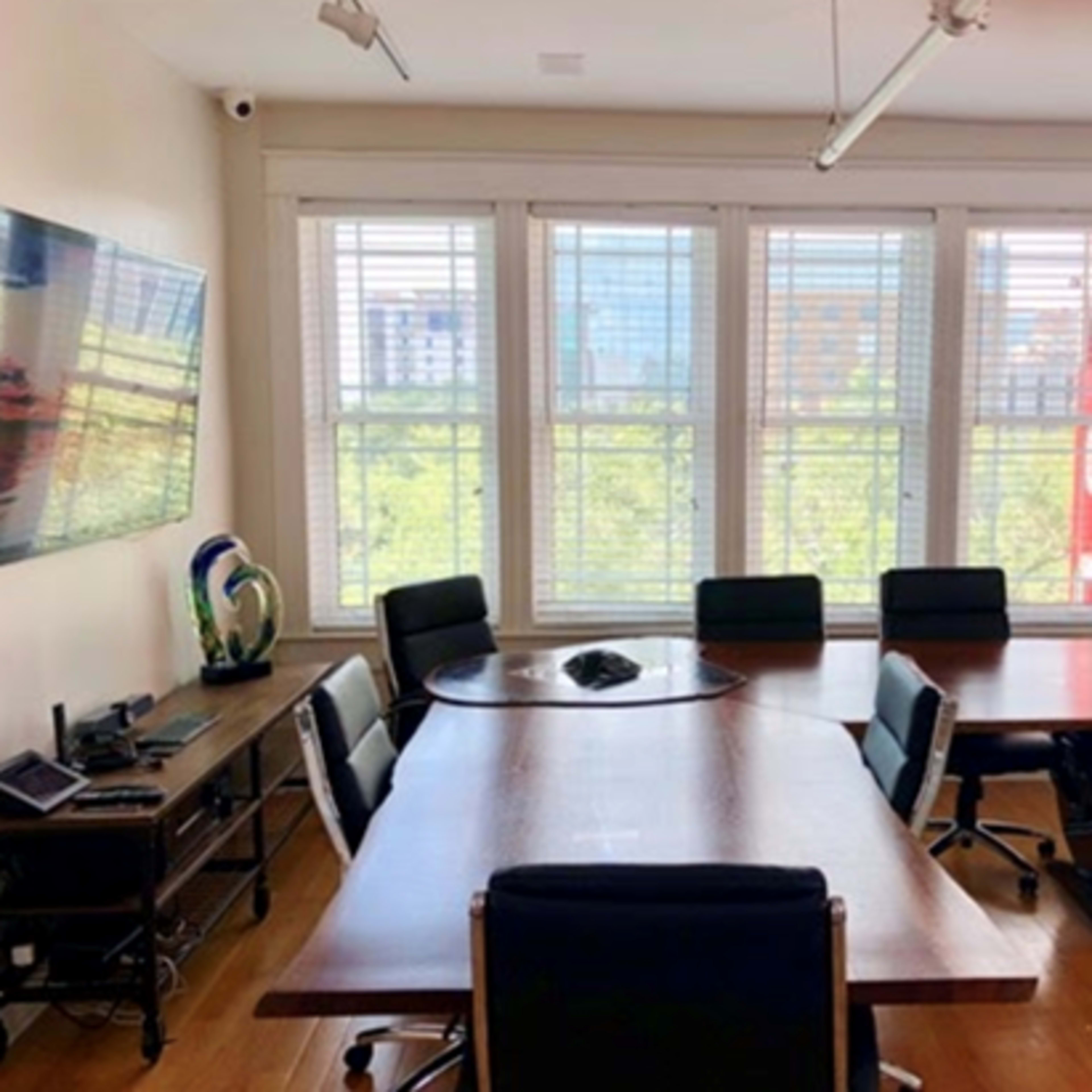A conference room features a large wooden table surrounded by chairs, with a television on the wall and large windows overlooking a greenery-filled view.