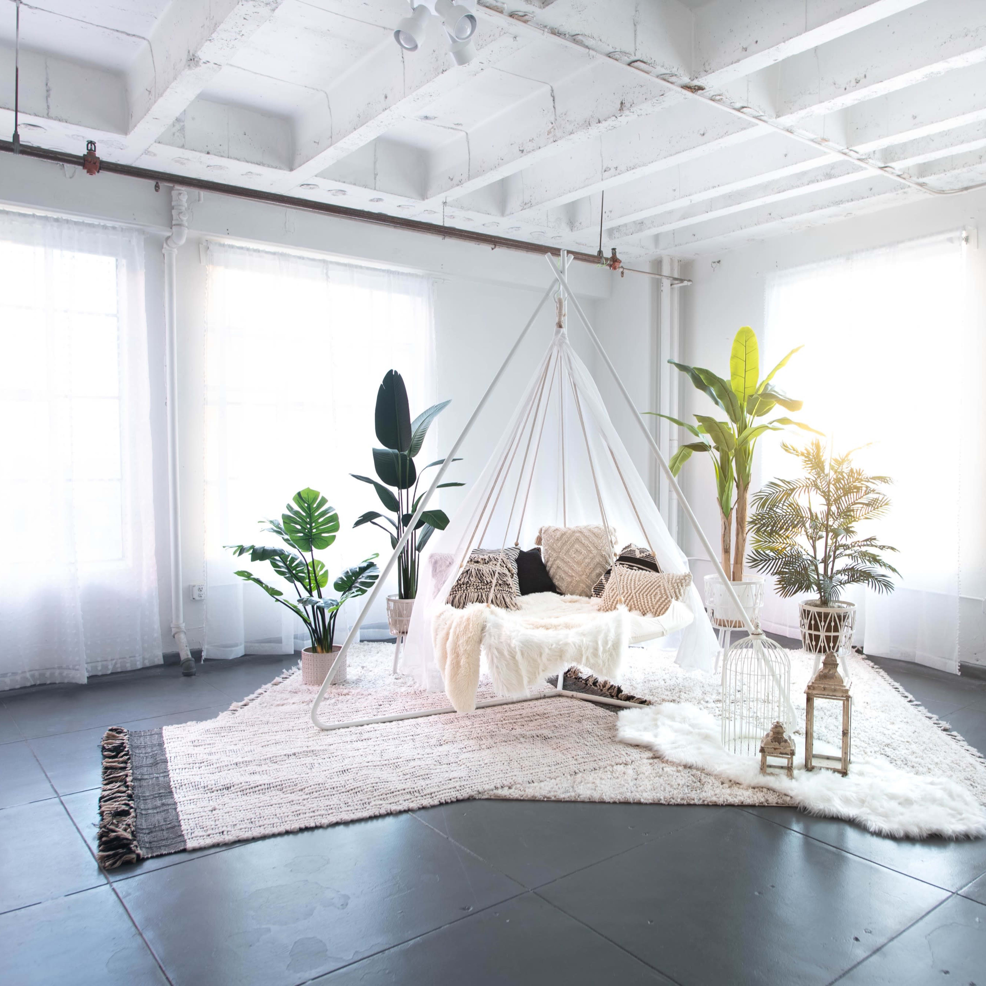 A bright, minimalistic interior space featuring a hanging chair surrounded by various plants on a large, textured rug.