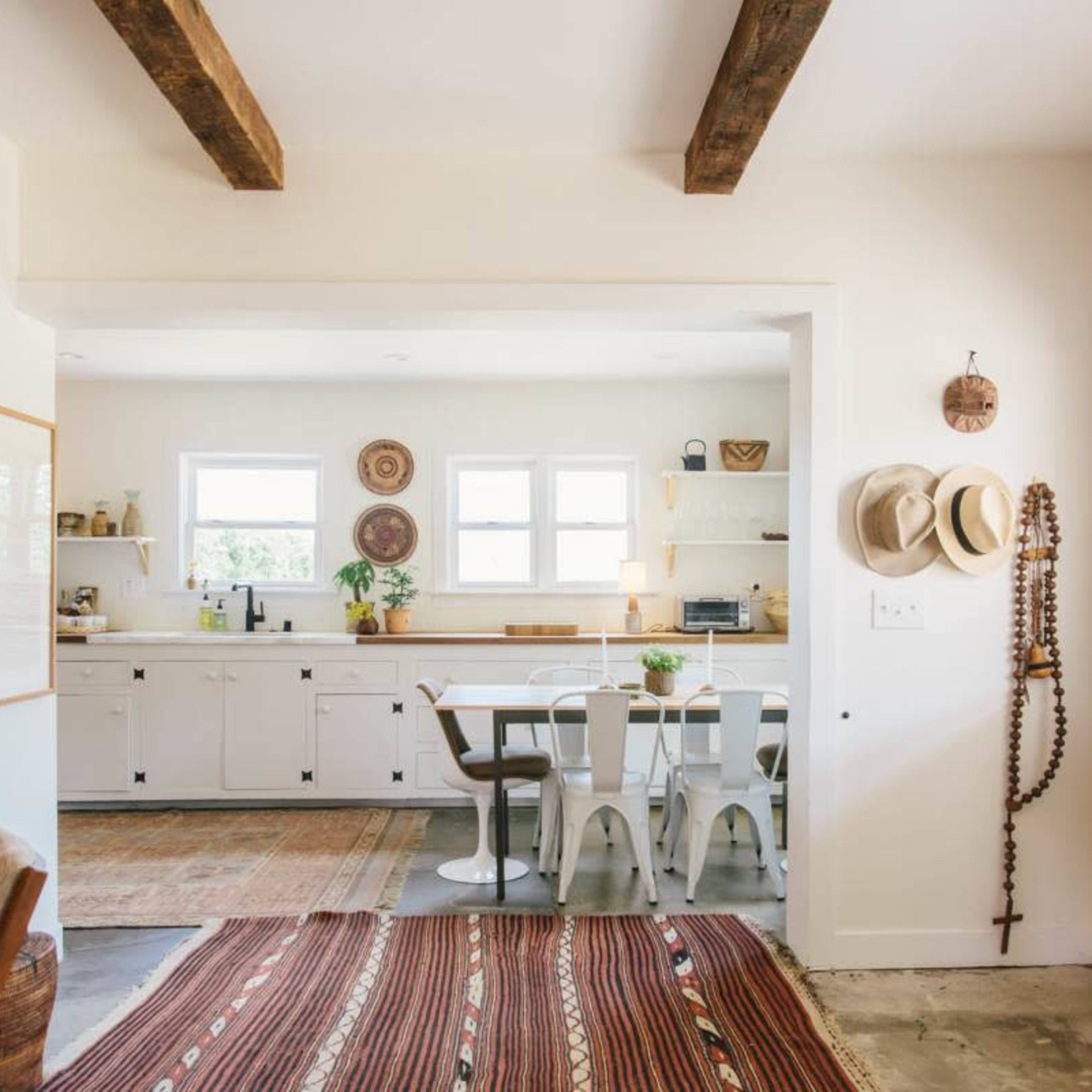 A bright open-concept living space features a kitchen and dining area with wooden beams, a patterned rug, and various decorative items.