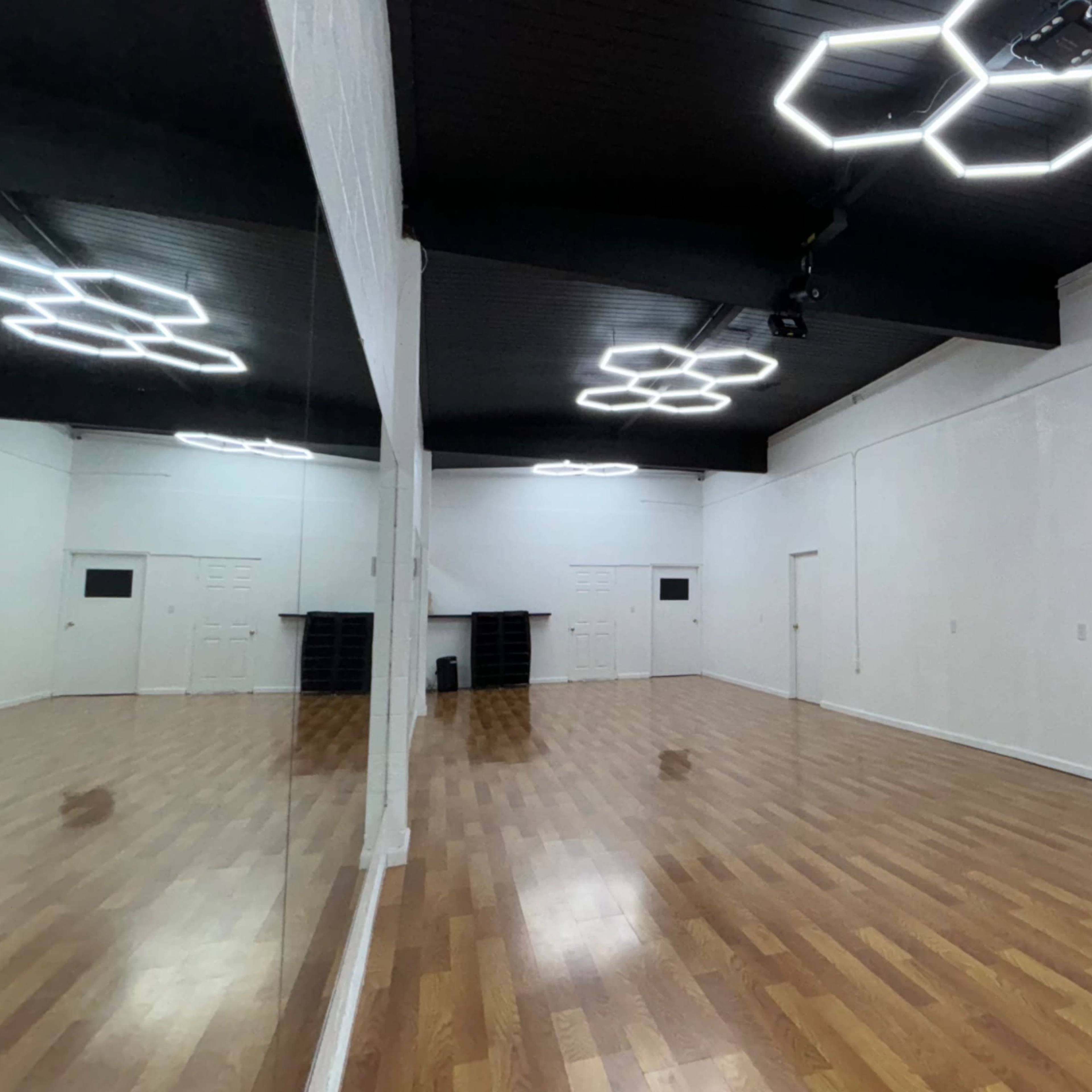A spacious, empty dance studio with wooden flooring, white walls, and modern light fixtures on the ceiling.