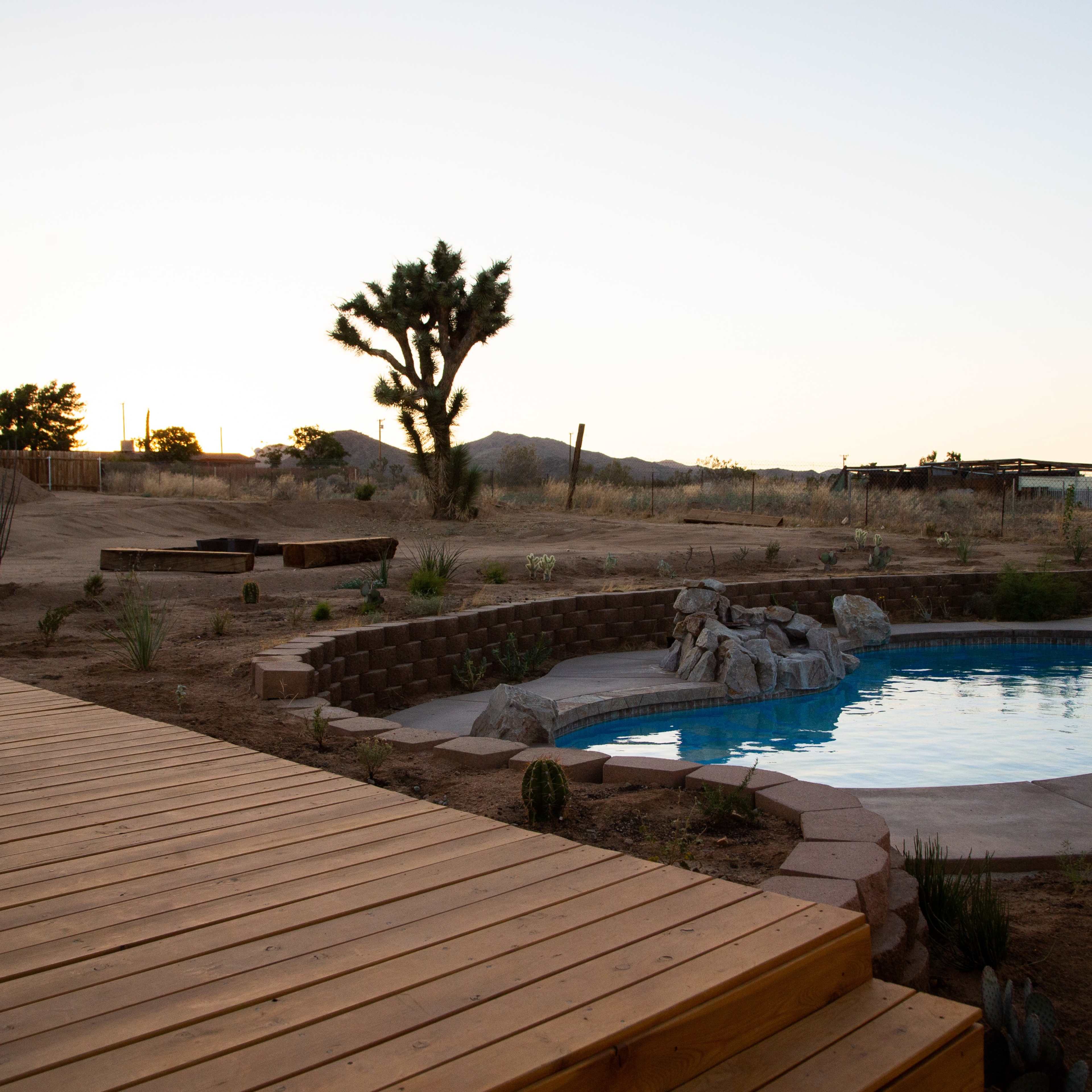 A wooden deck overlooks a swimming pool surrounded by stone landscaping and shrubs in a desert landscape at sunset.