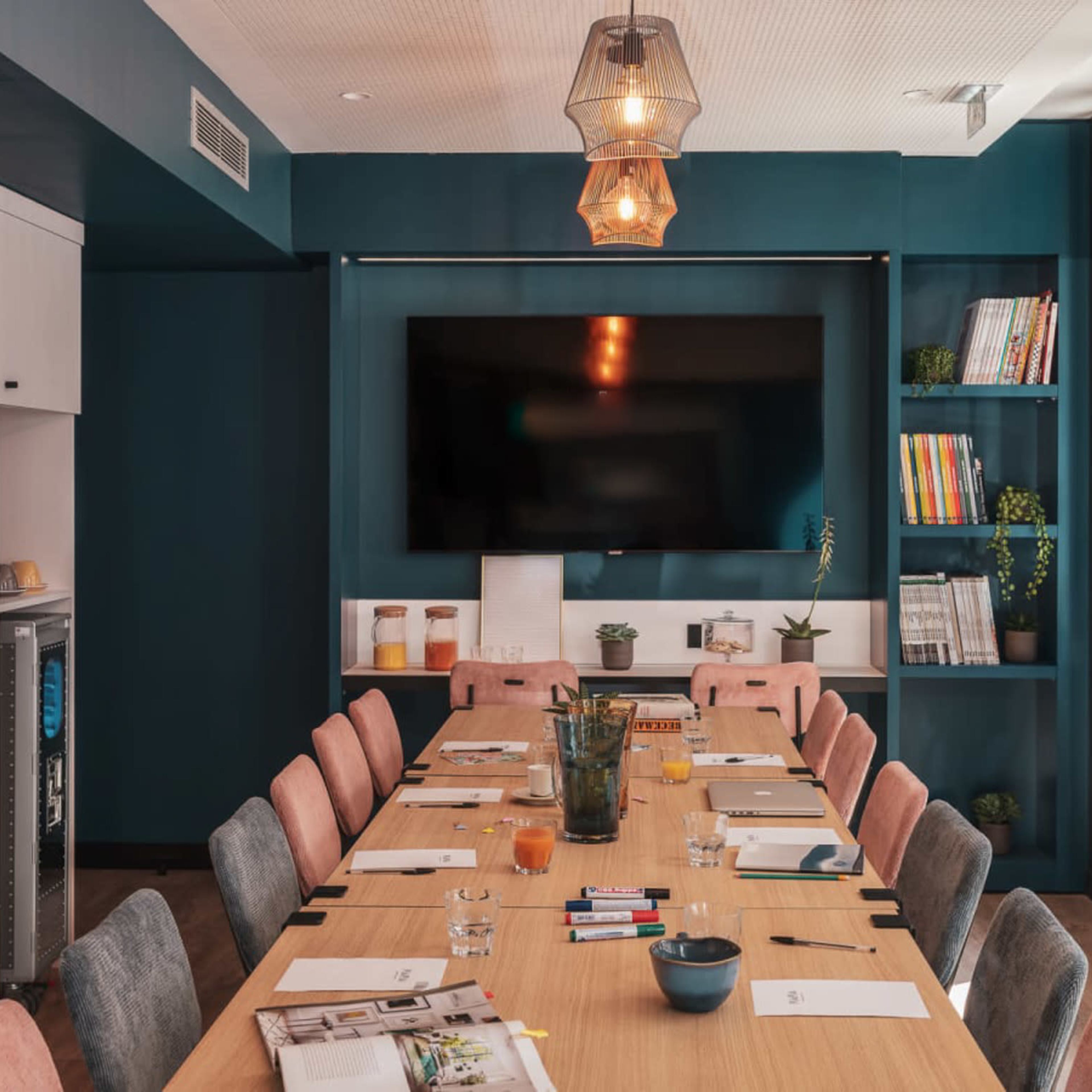 A long wooden conference table with chairs is set up in a room featuring a large television and shelves filled with books and plants.
