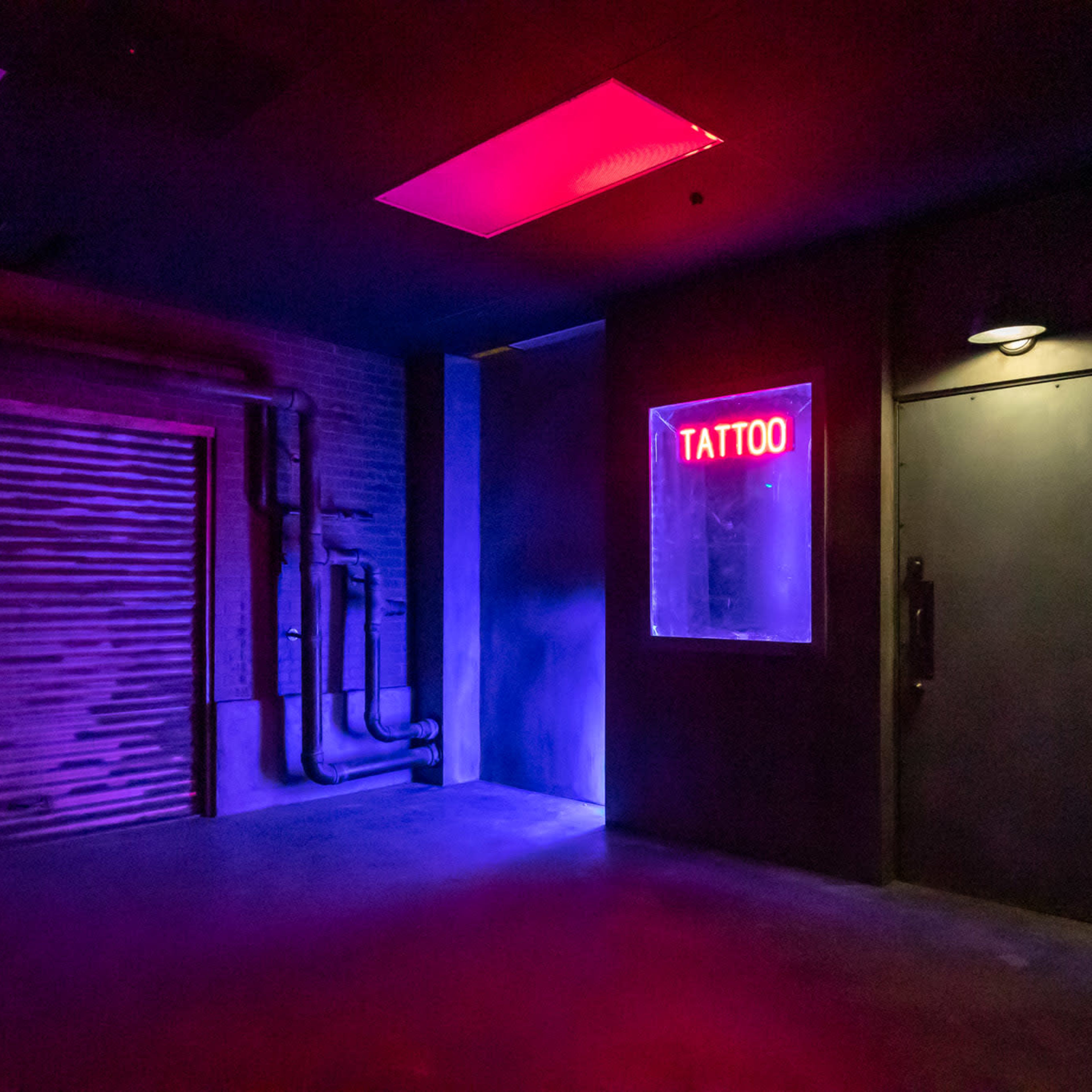An underground room illuminated by purple and red lights, featuring a closed door with a "TATTOO" sign and exposed pipes along the wall.