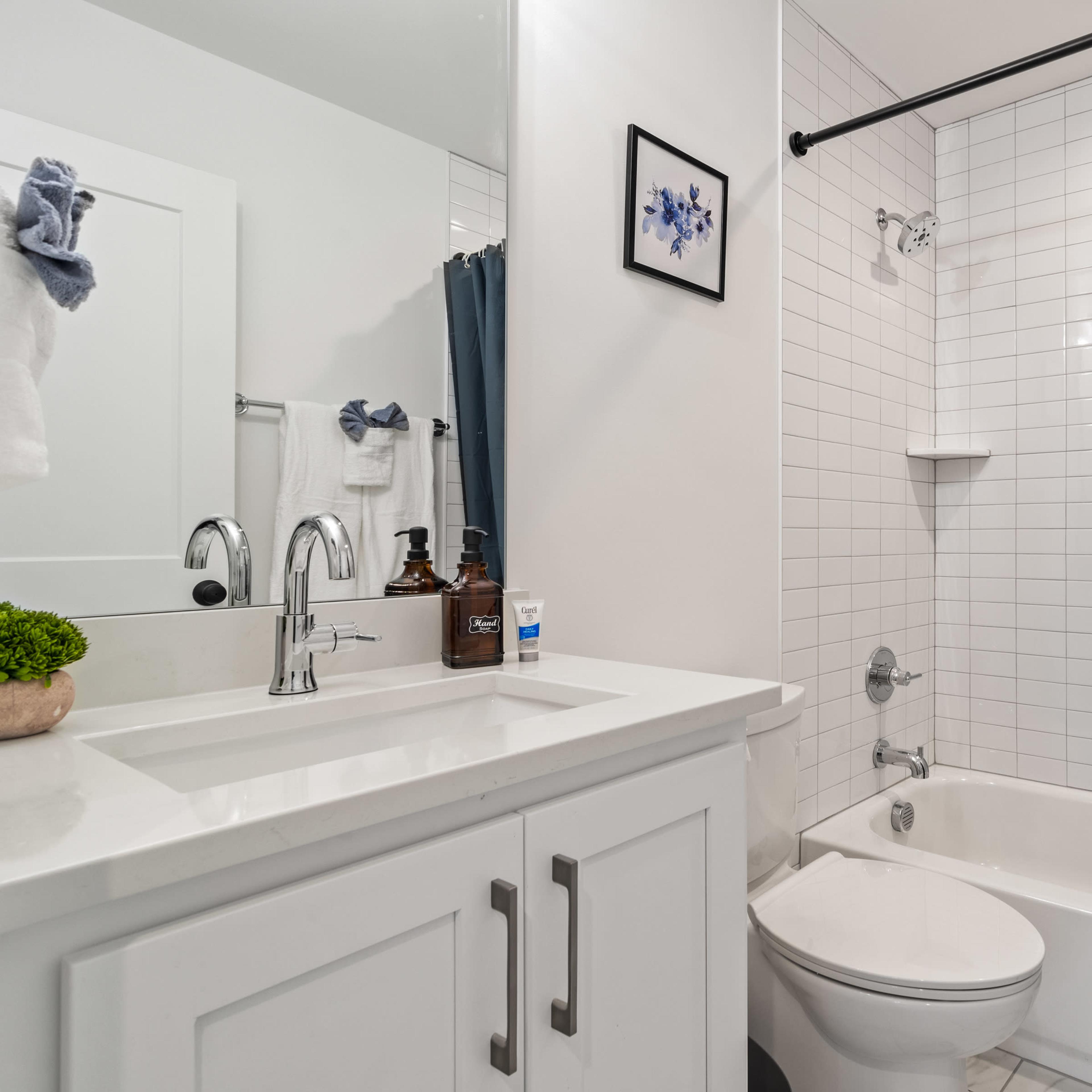 A modern bathroom featuring a white tiled tub and shower area, a sink with a mirror, and a small plant on the counter.