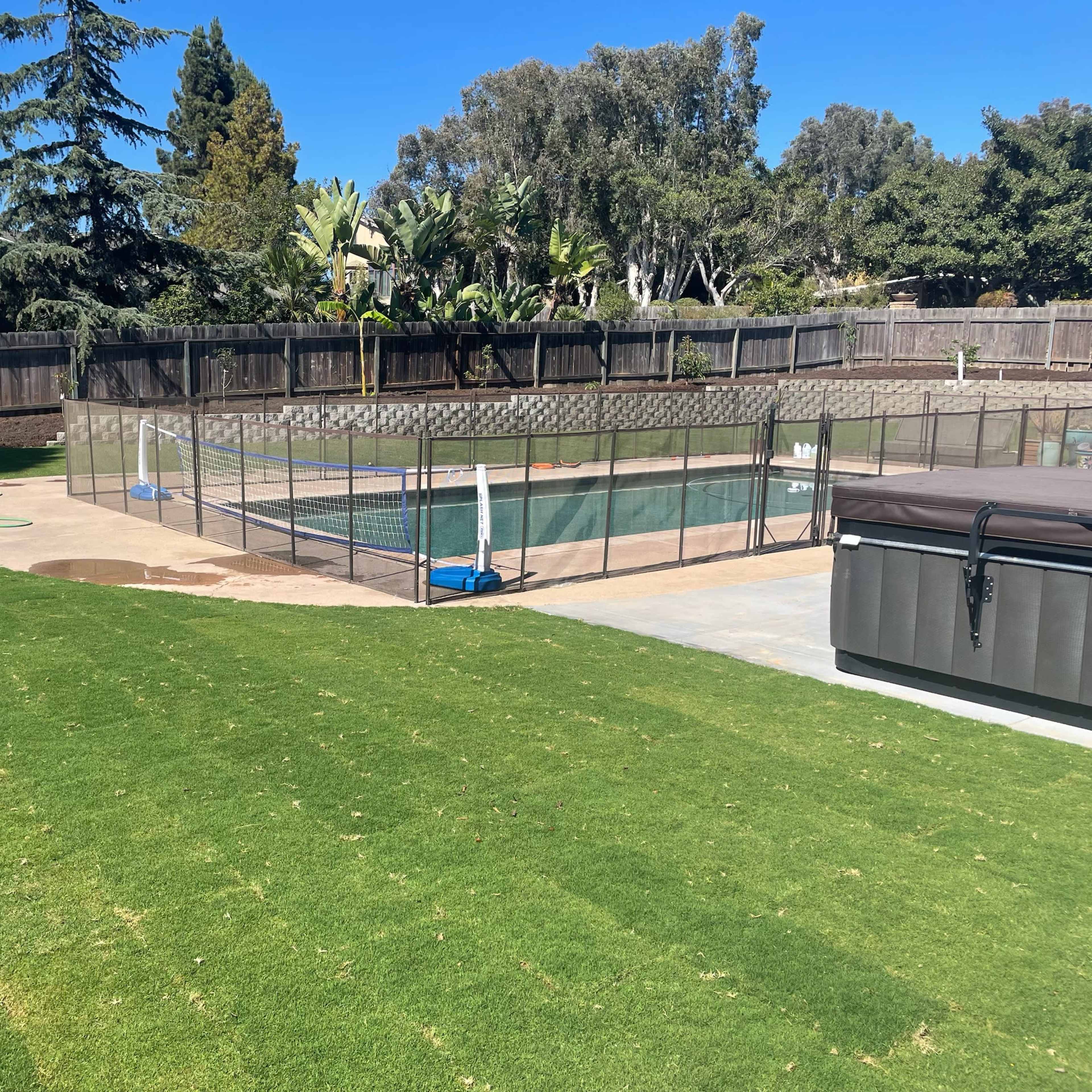 A backyard with a green lawn, a fenced swimming pool area, and a hot tub.