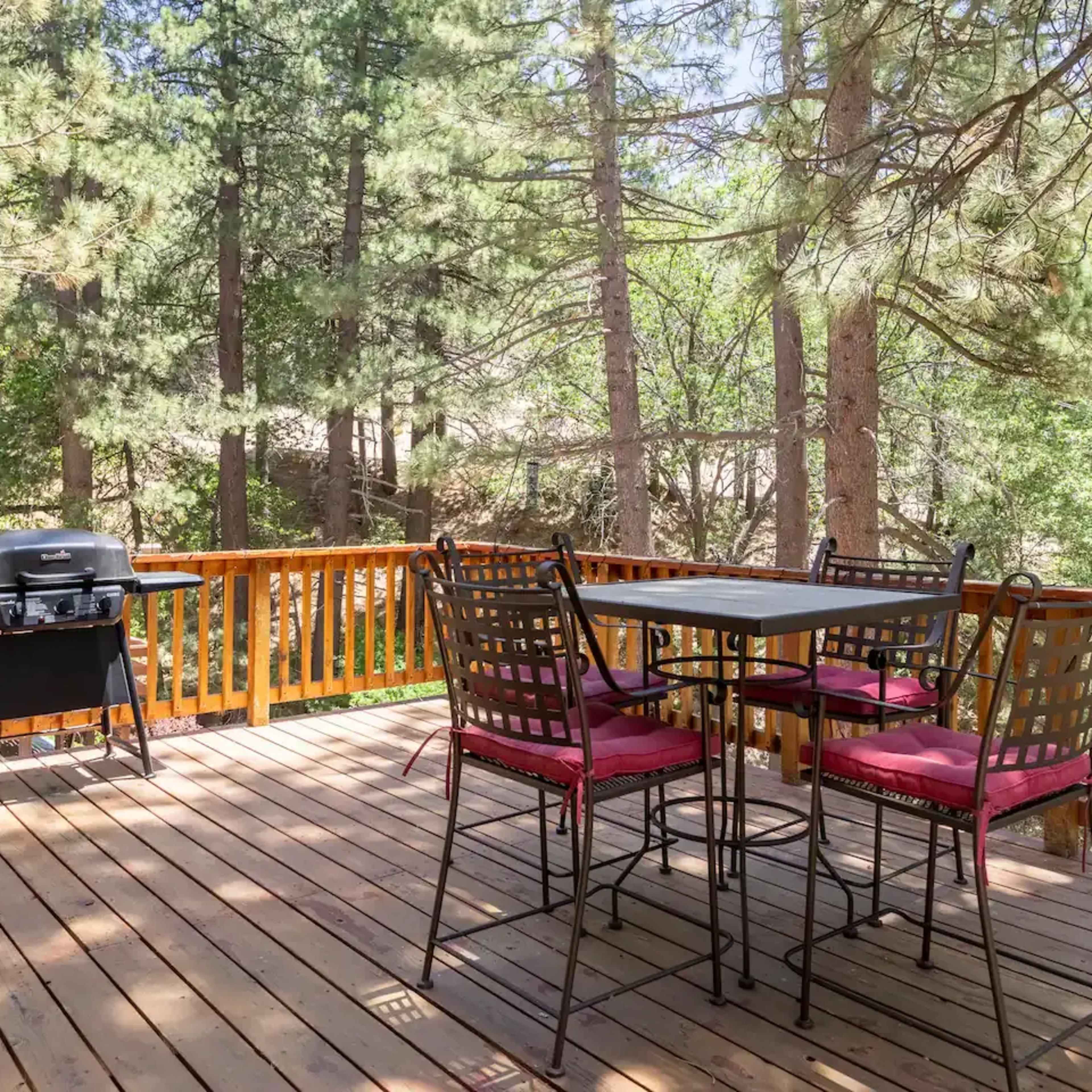 A wooden deck surrounded by trees features a table with four chairs and a grill nearby.