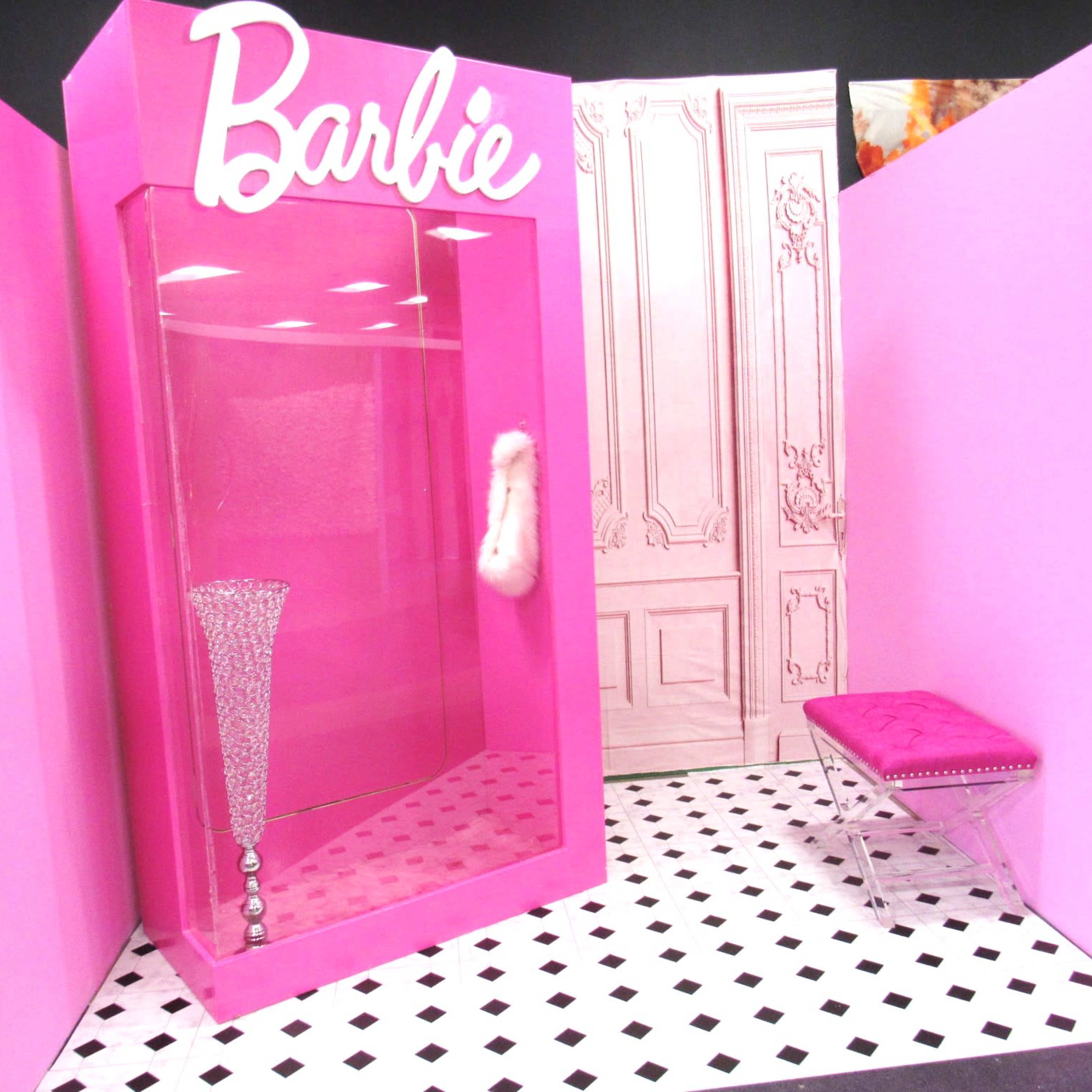 A pink display featuring a glass case labeled "Barbie," with a crystal vase, a pink fur item, and a clear acrylic bench in a room with a patterned floor.