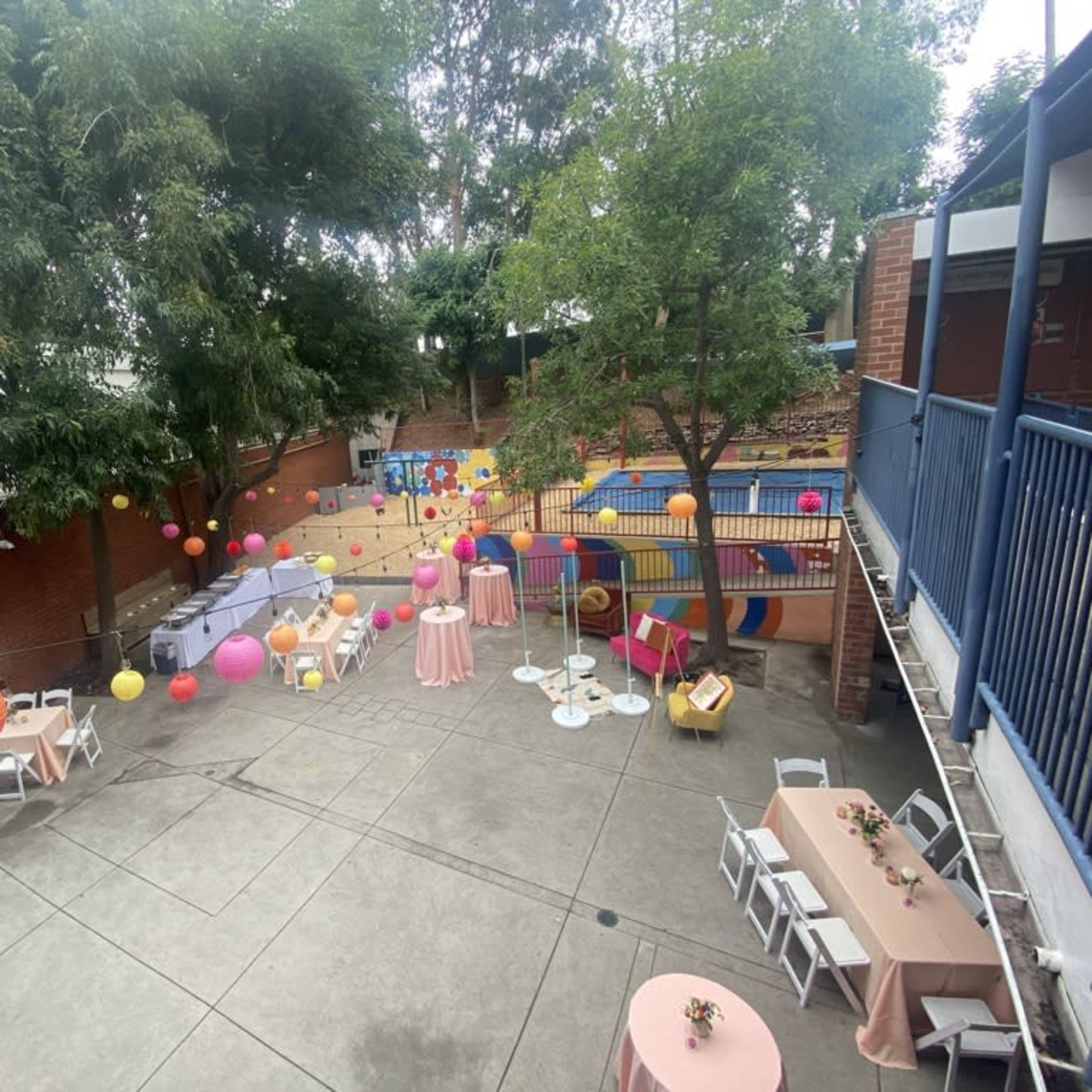 An outdoor event space with tables dressed in pink linens, colorful lanterns hanging from trees, and a playground area in the background.