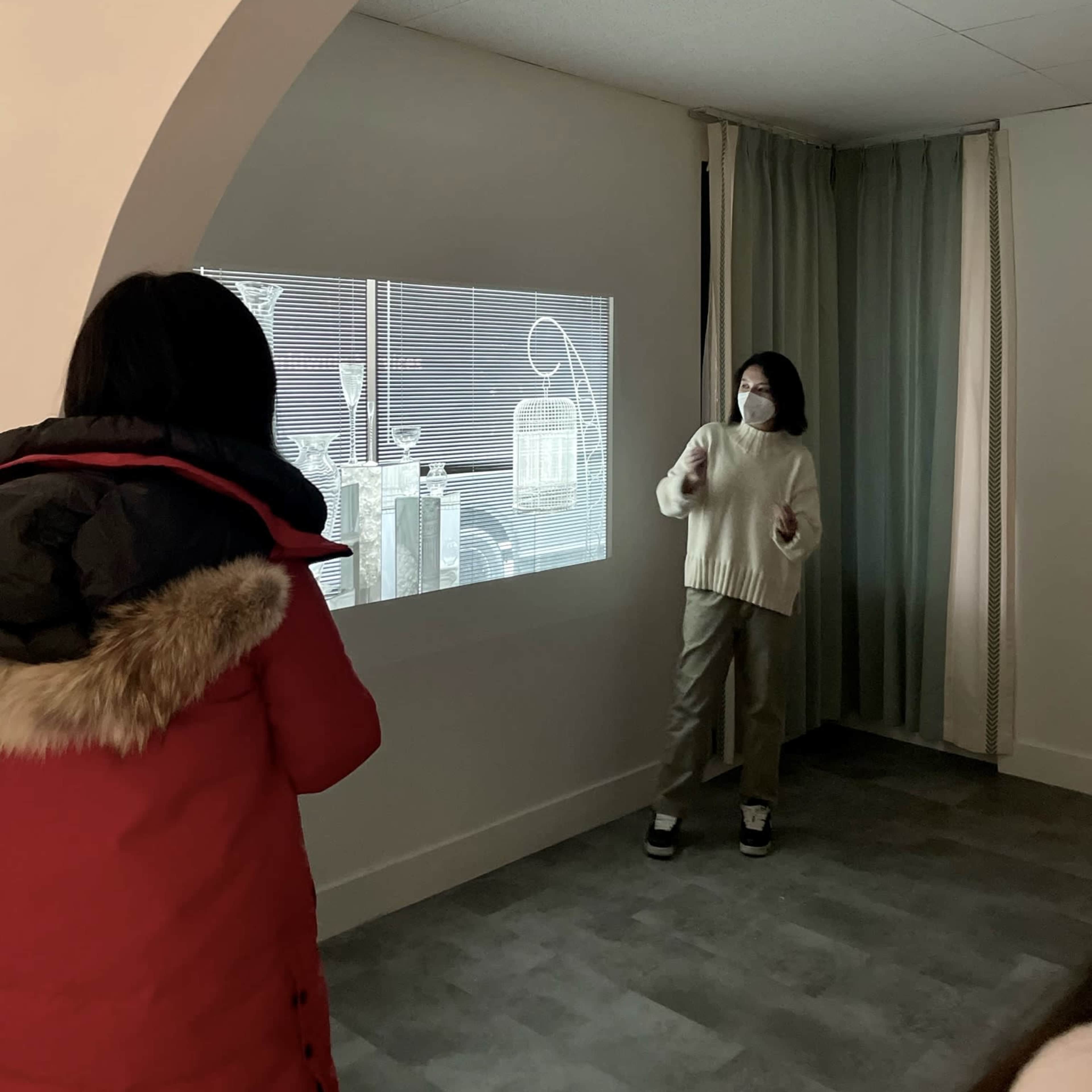 A person in a white sweater presents in front of a projected image while another individual in a red coat listens attentively.