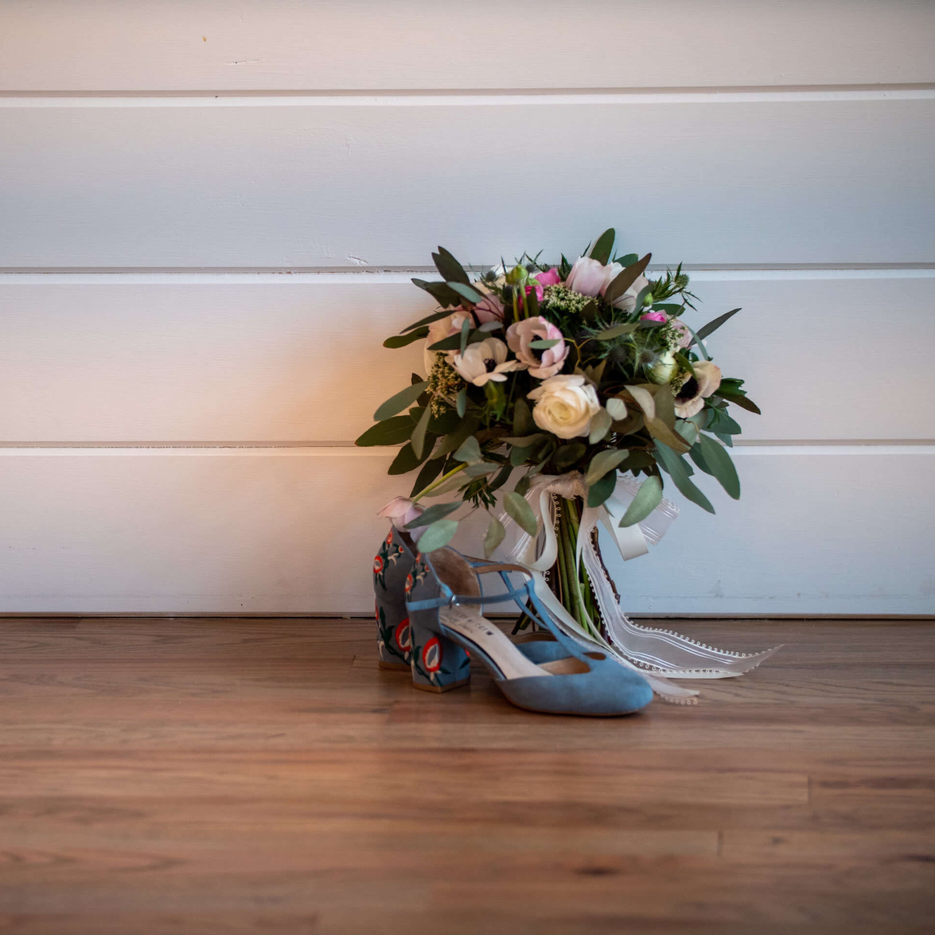 A pair of blue shoes sits next to a floral bouquet against a white wooden backdrop.