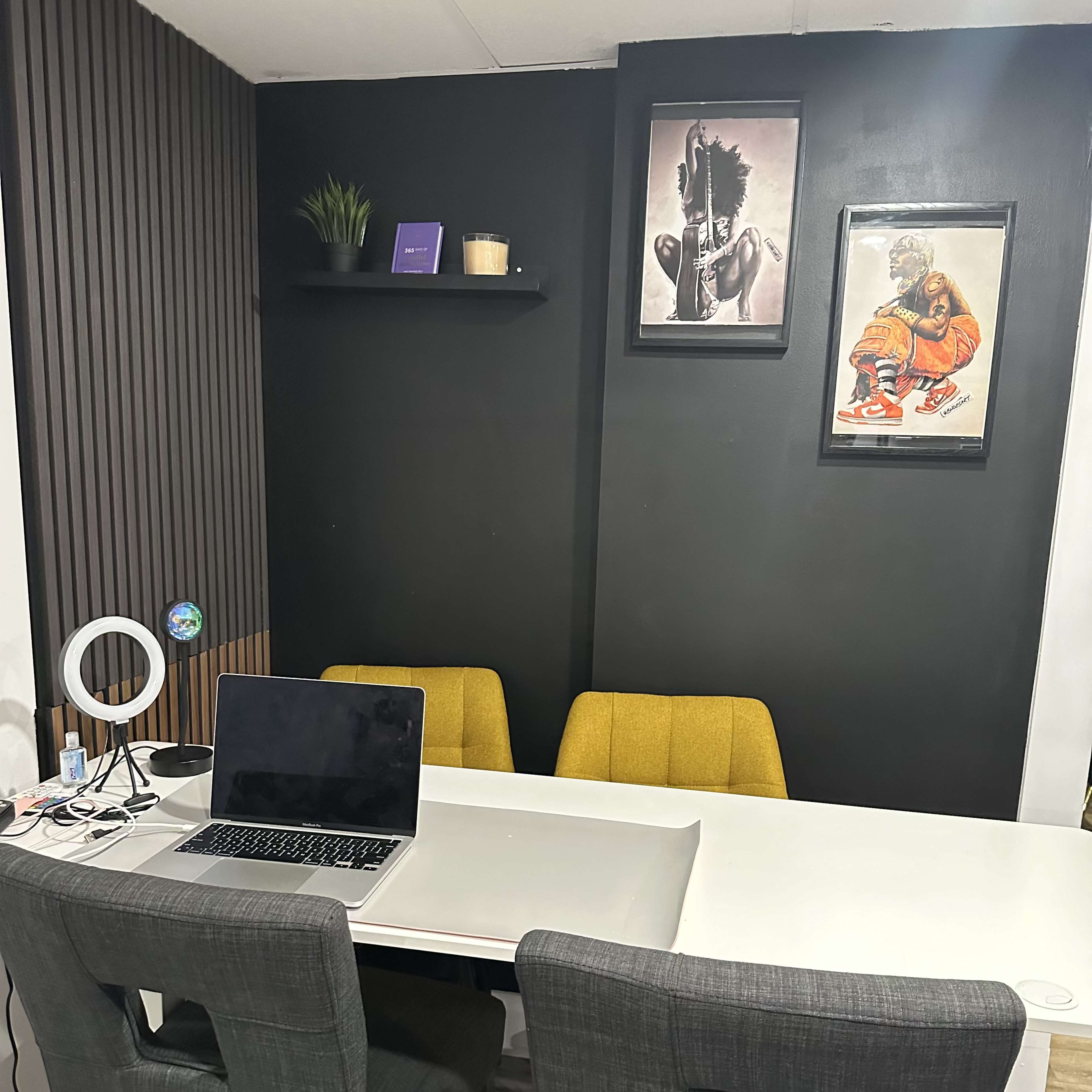 A modern office space with a white table, four chairs, a laptop, and two framed artworks on a black wall.