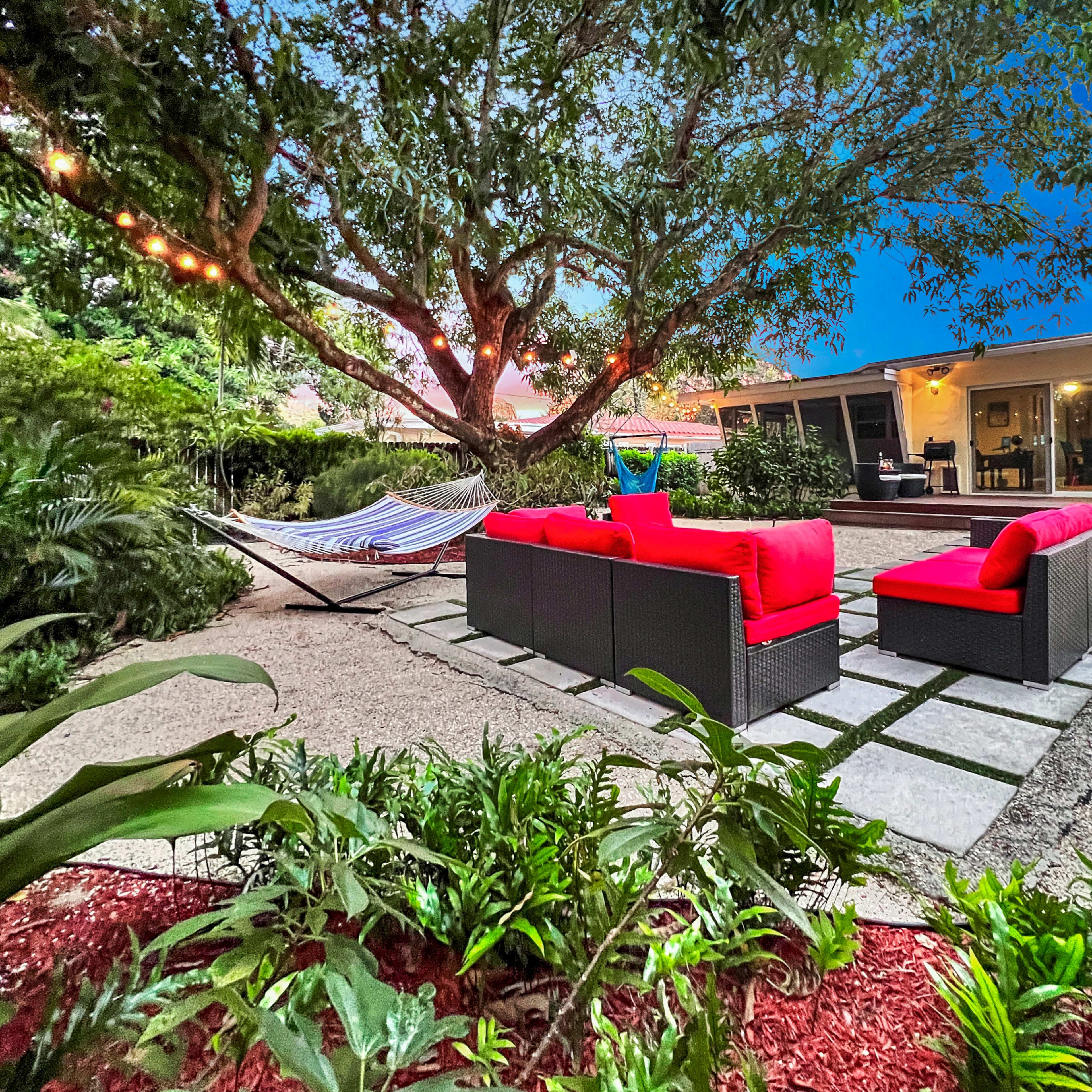 A cozy outdoor seating area features red cushions on black wicker furniture arranged around a stone path, with a hammock and garden greenery nearby.