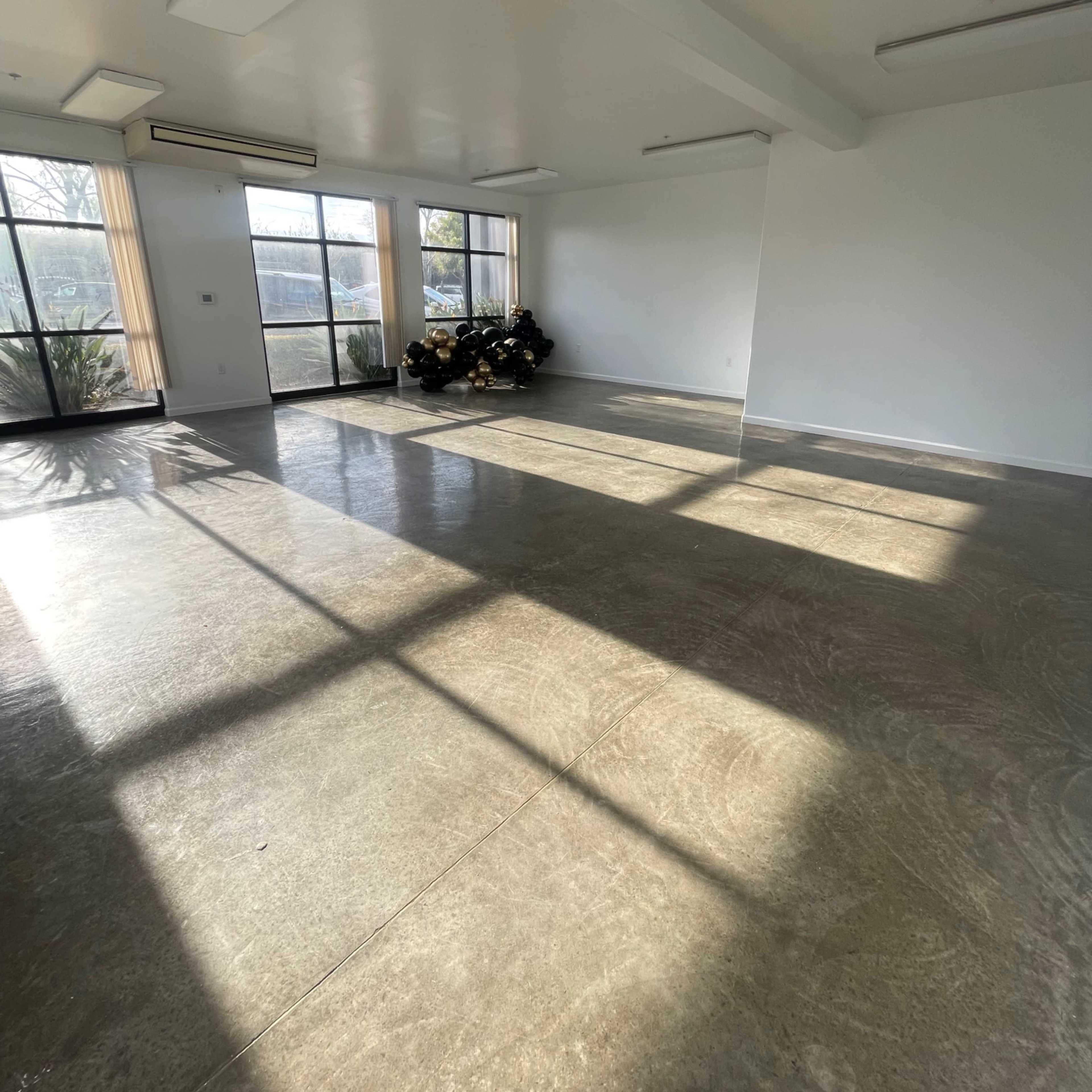 A spacious, empty room with large windows allowing sunlight to create distinct shadows on the polished floor.